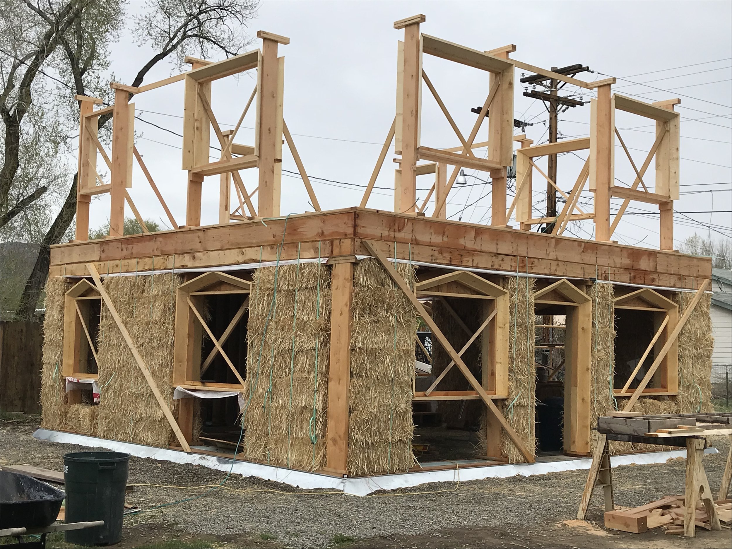 Construction site with wooden framing and hay bales for a building, with trees and utility poles in the background.