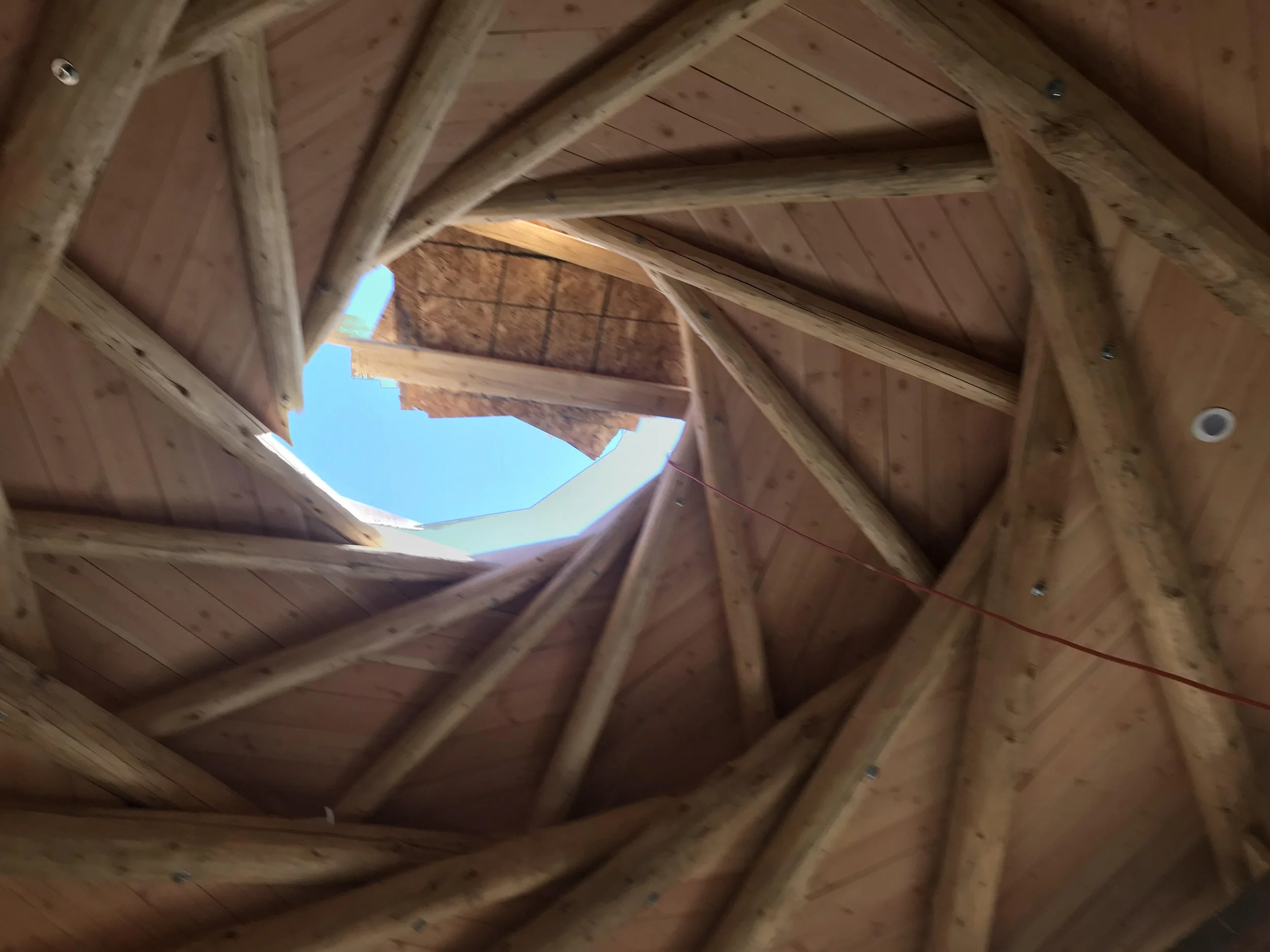 The photo shows the inside of the roof of a house made of wooden beams and panels, with a circular opening in the roof revealing the blue sky outside.