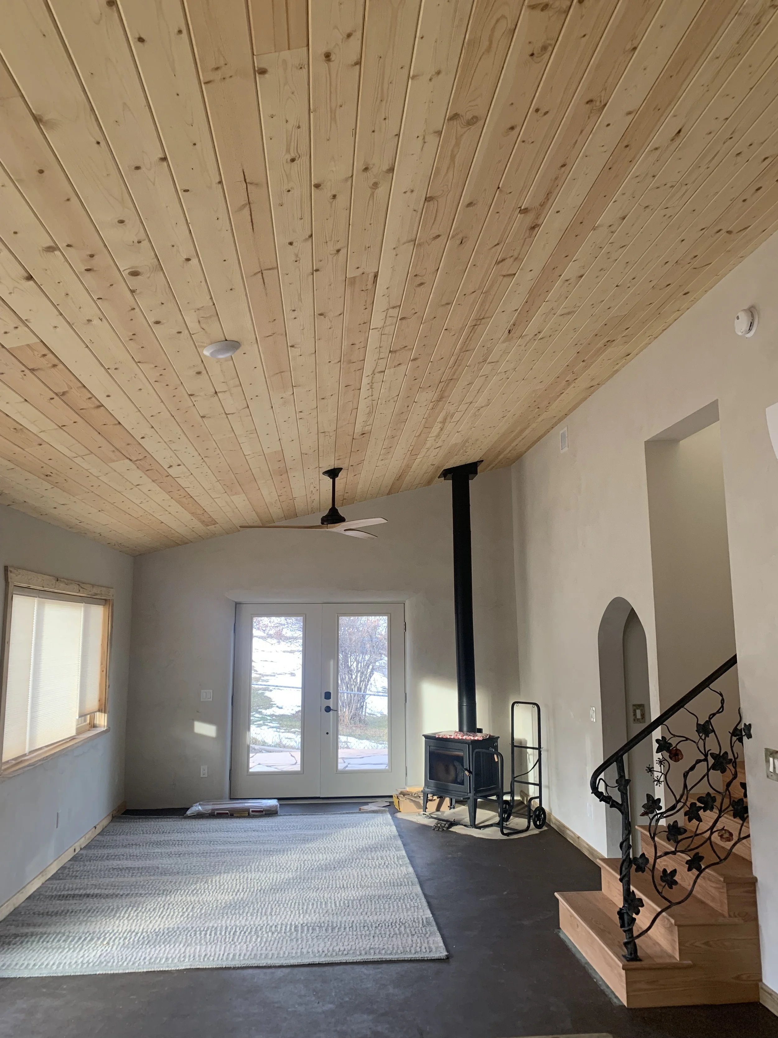 Living room with a wooden ceiling, a ceiling fan, a door with glass panels, a window, a small heating stove, and a staircase with a decorative black iron railing.