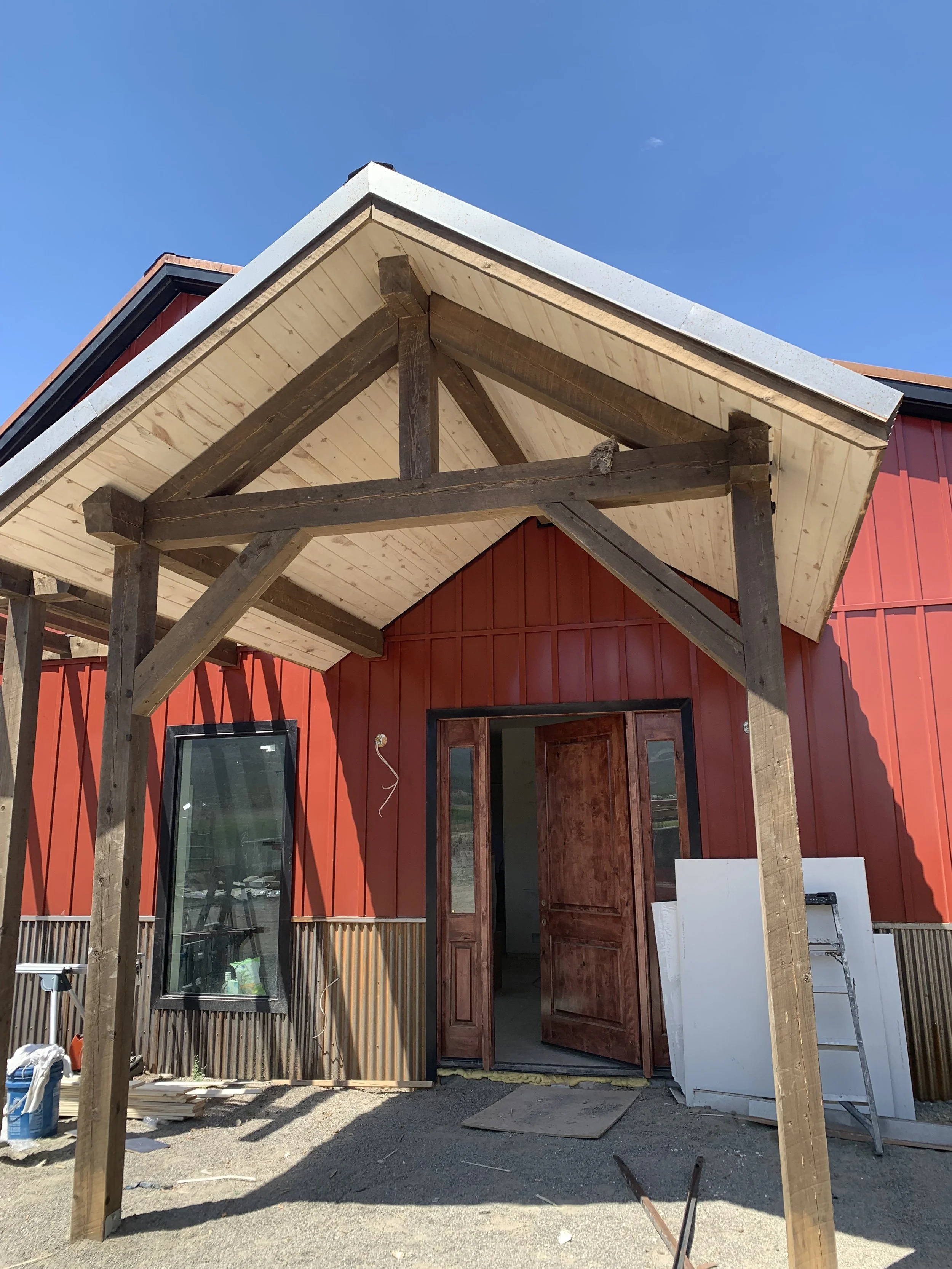 Construction of a building entrance with a wooden roof structure and a red wall, open door, construction tools and materials nearby under a clear blue sky.