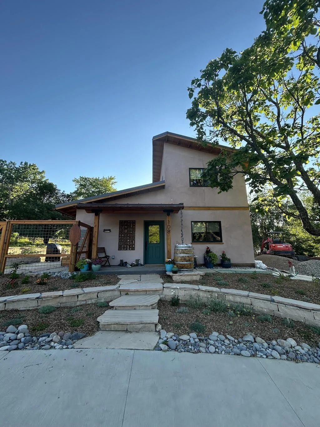 A modern two-story house with a small front porch, surrounded by a landscaped yard with rocks and plants. There are several potted plants near the entrance, and a barrel with a hanging chain. A red tractor is visible on the right side of the yard, and a large tree with green leaves is on the right side of the image. The sky is clear and blue.