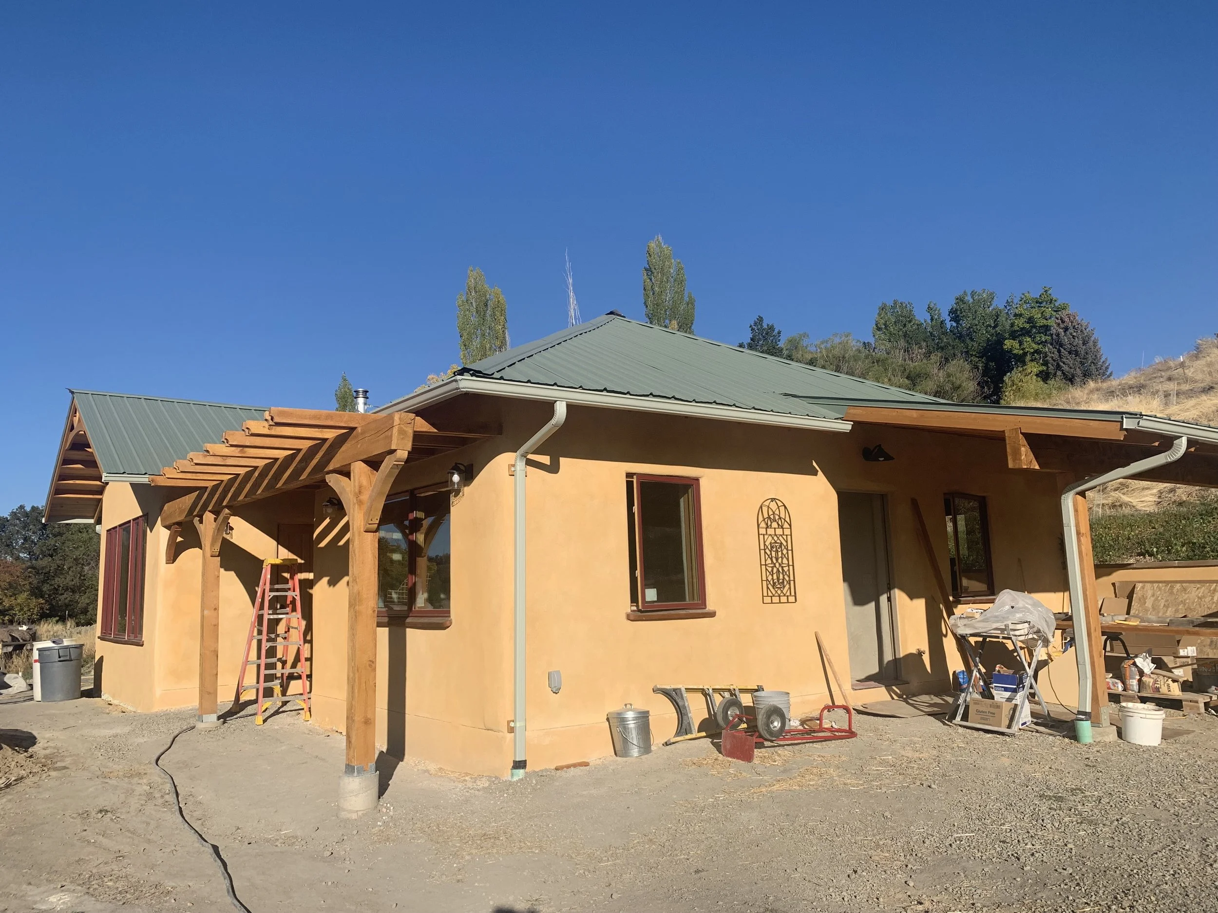 A house under construction with beige walls, a green metal roof, and wooden beams supporting a porch area. Construction tools and materials are scattered around the site.