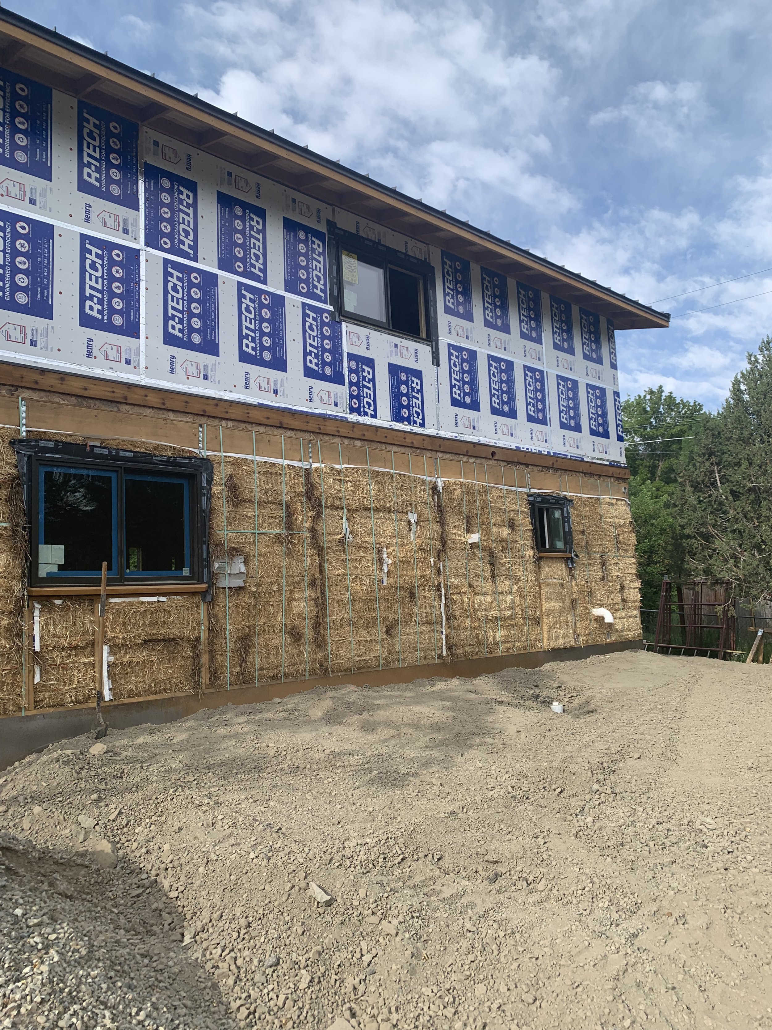 A house under construction with straw insulation on the exterior walls, two small windows, and a partially completed ground area.