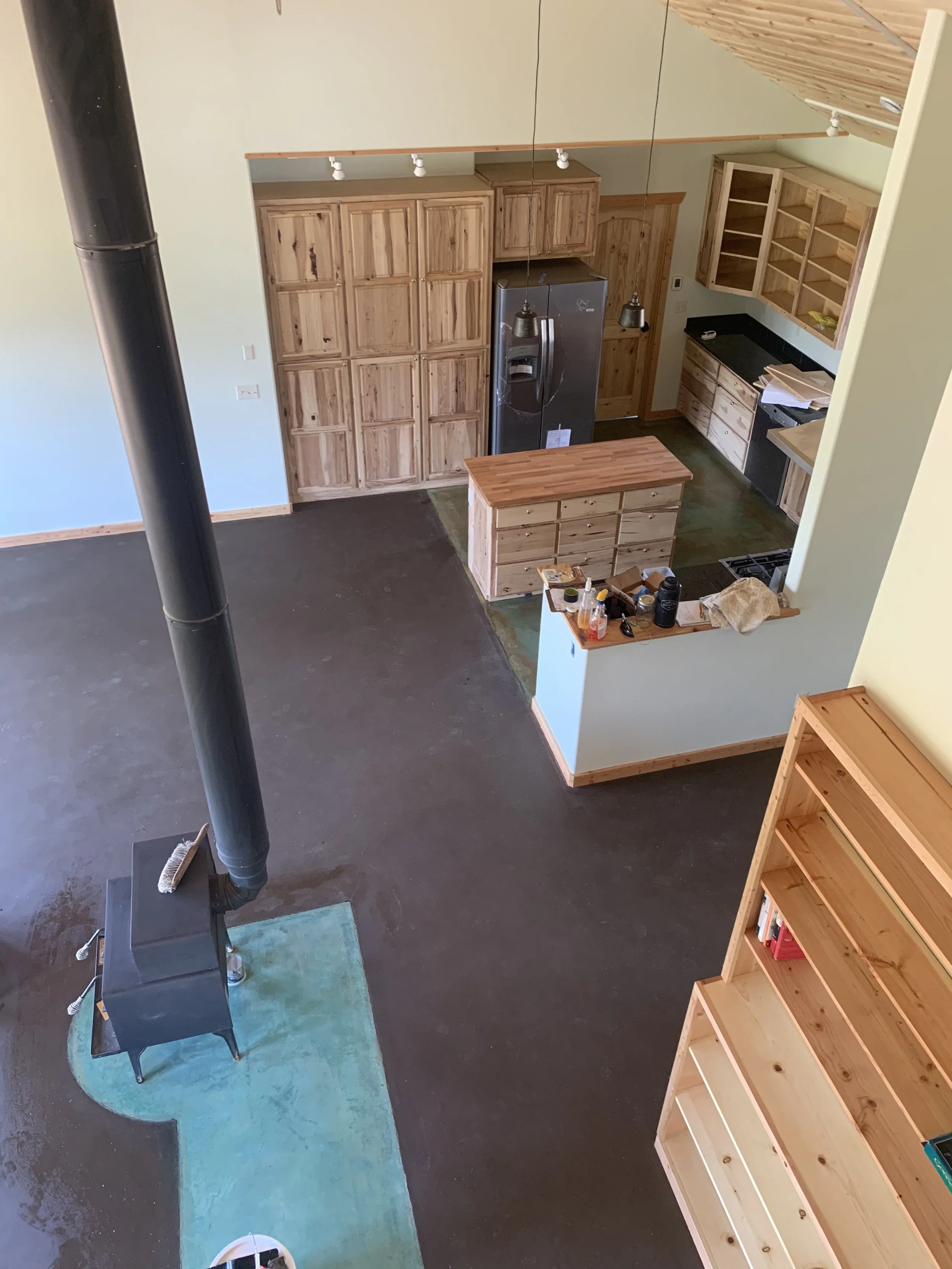 A kitchen under construction with wooden cabinets, a black refrigerator, a small island, and open shelving. A wood stove pipe extends from the left. The ceiling is sloped with wood paneling. There are work supplies on a small counter.
