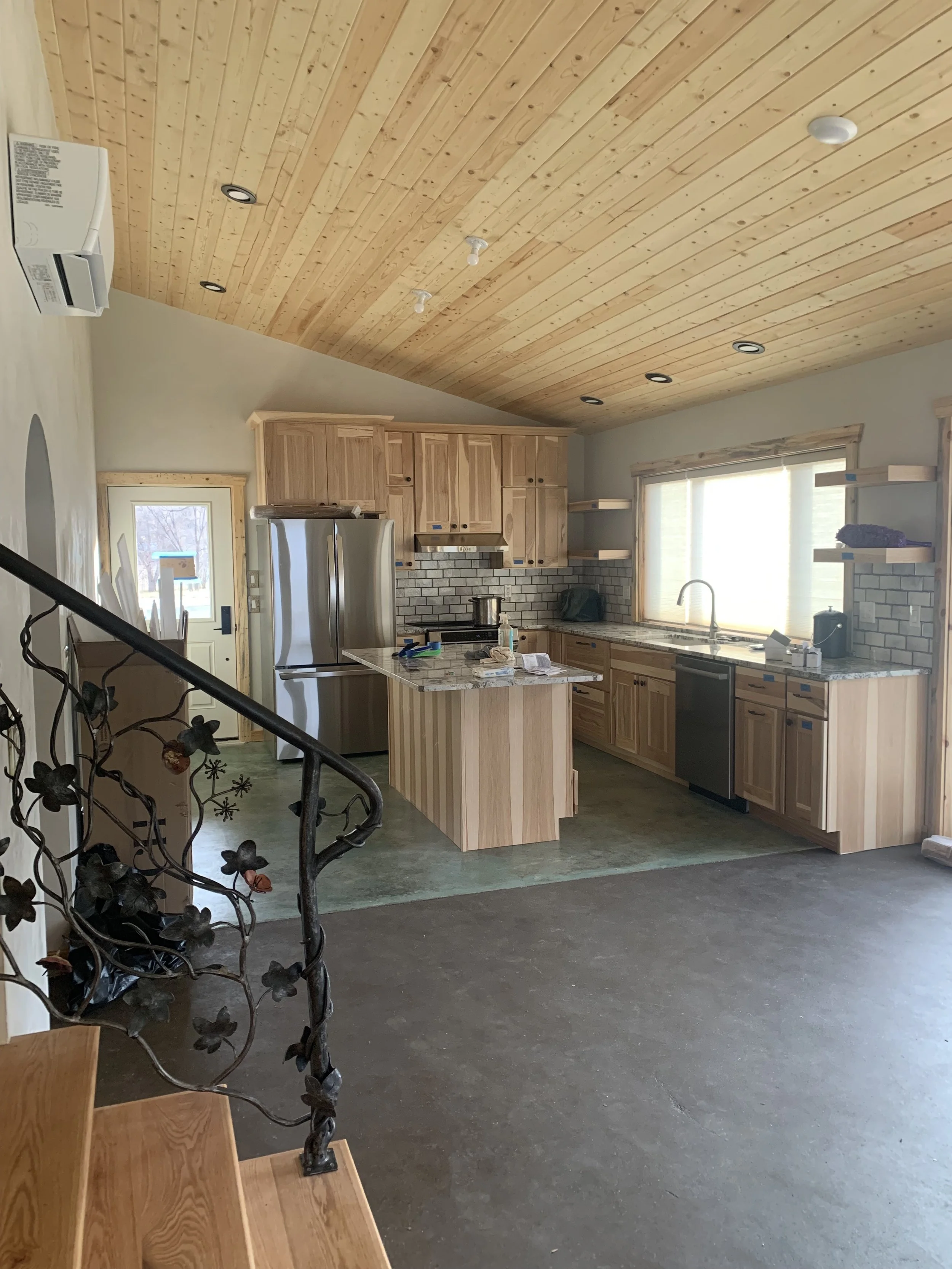 Interior of a kitchen with unfinished cabinets, a granite countertop, a stainless steel refrigerator, and a window with blinds. The ceiling is made of wood paneling, and there are recessed lights installed. Some construction tools and materials are o
