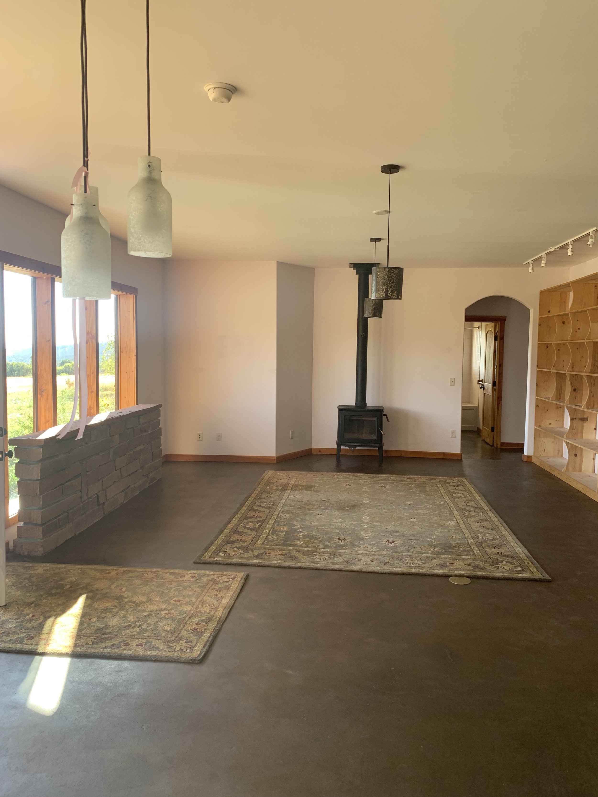 Interior of a room with a wood stove, hanging light fixtures, a patterned rug, and large windows showing an outdoor landscape.