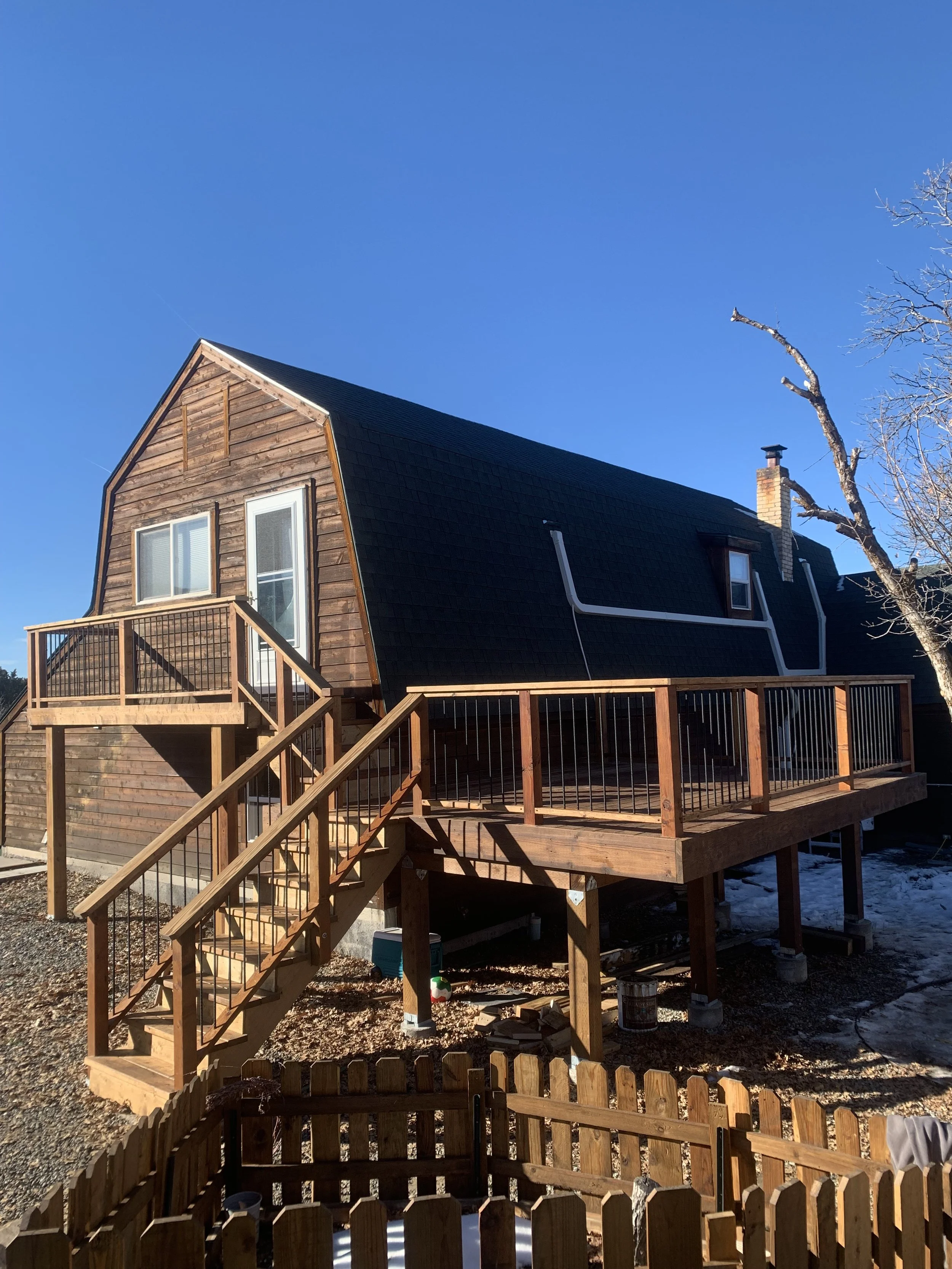 The image shows a house with a sloped black roof and wooden siding, under construction with a wooden deck and stairs, set against a clear blue sky with a leafless tree to the right.
