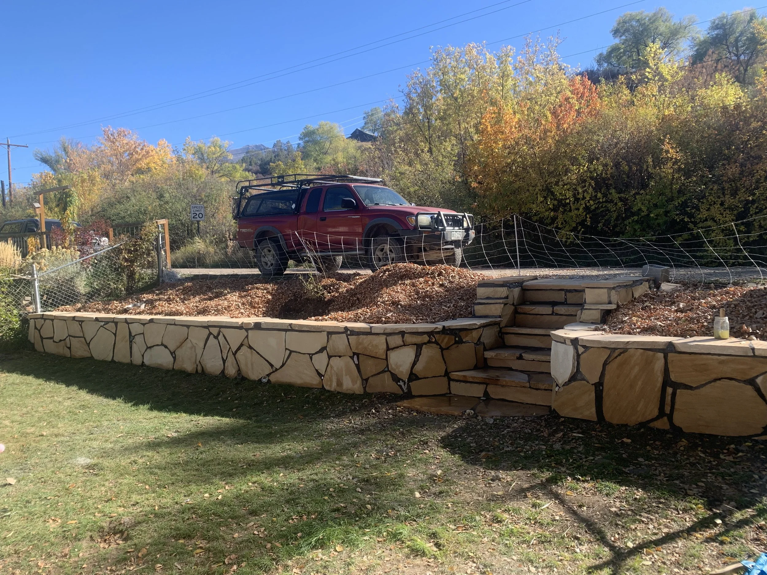 A red SUV with a roof rack parked on a dirt area surrounded by trees with colorful autumn leaves, a stone wall, and a wooden fence, with mountains and power lines in the background.