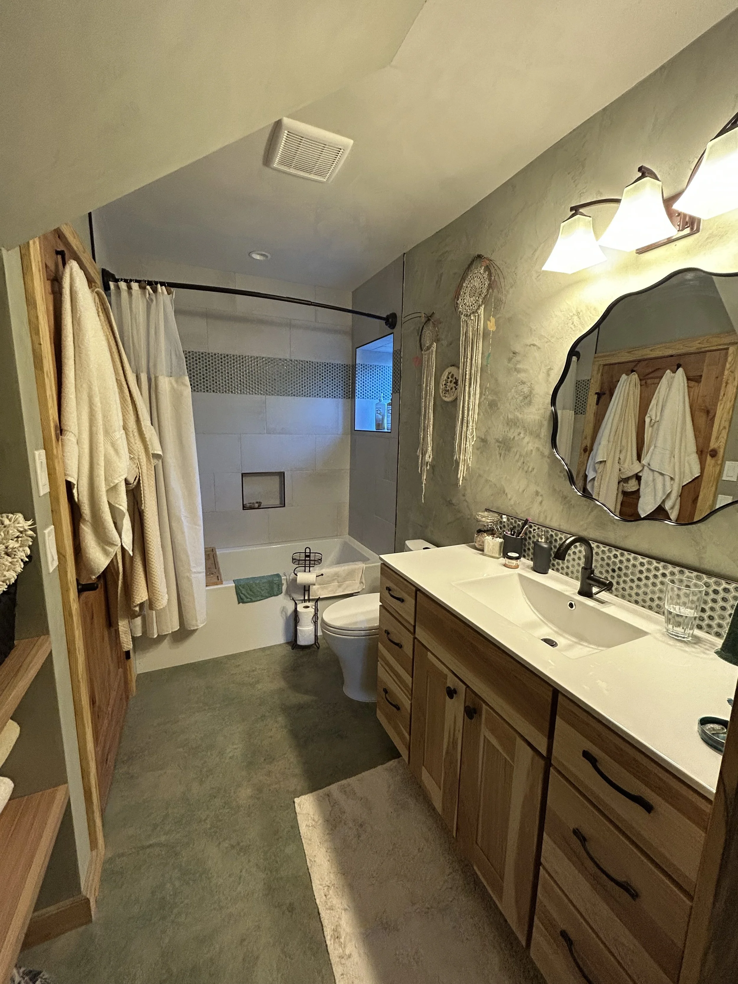 A bathroom with a wooden cabinet vanity, a large mirror, and wall-mounted light fixtures. There are towels hanging on hooks and a shower area with a curtain, a small window, and a bathtub.