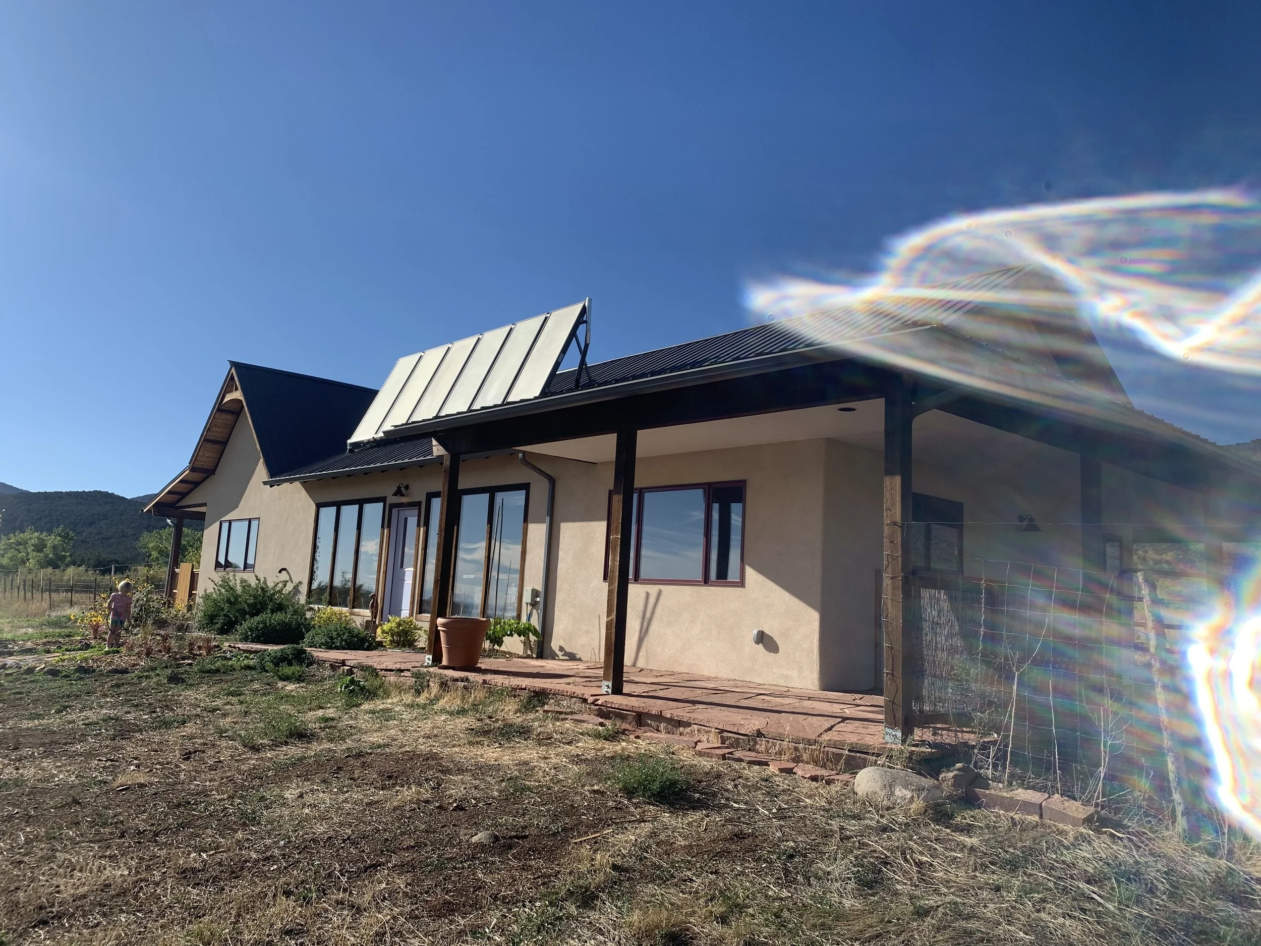 A house with a dark roof, beige walls, and several windows, set against a clear blue sky with minimal clouds. The house has a patio area with a potted plant and garden in the foreground. There is a lens flare on the right side of the image.