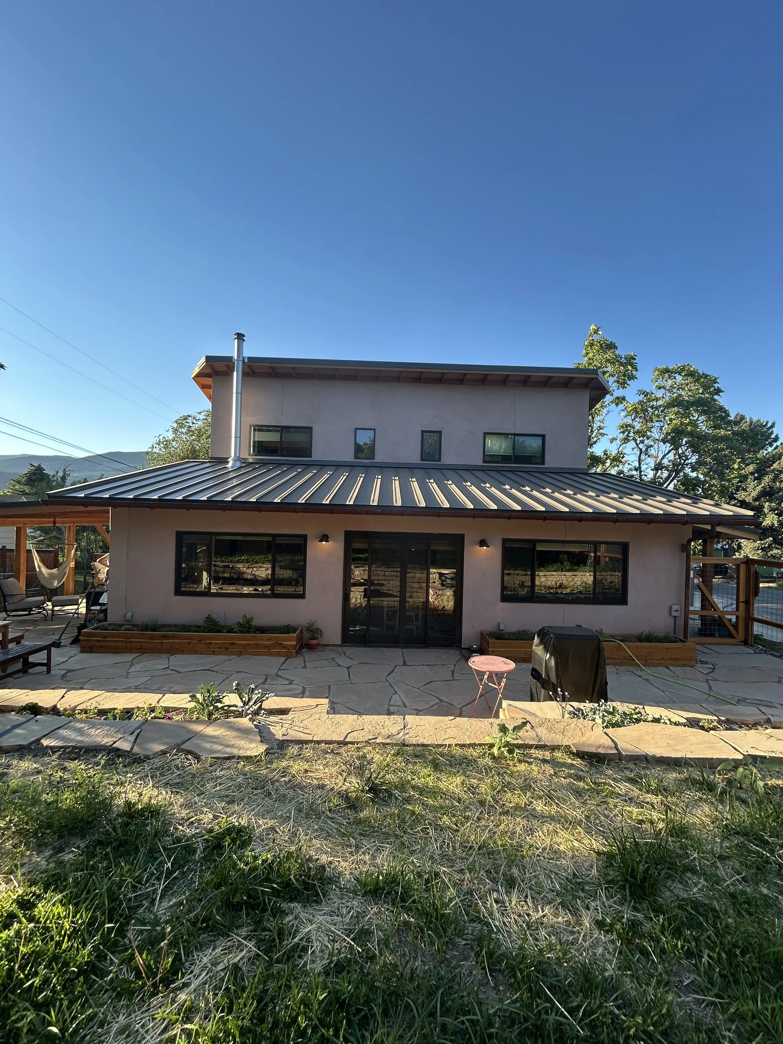 A two-story house with a metal roof, multiple windows, and a patio area with outdoor furniture, set against a backdrop of trees and mountains under a clear blue sky.