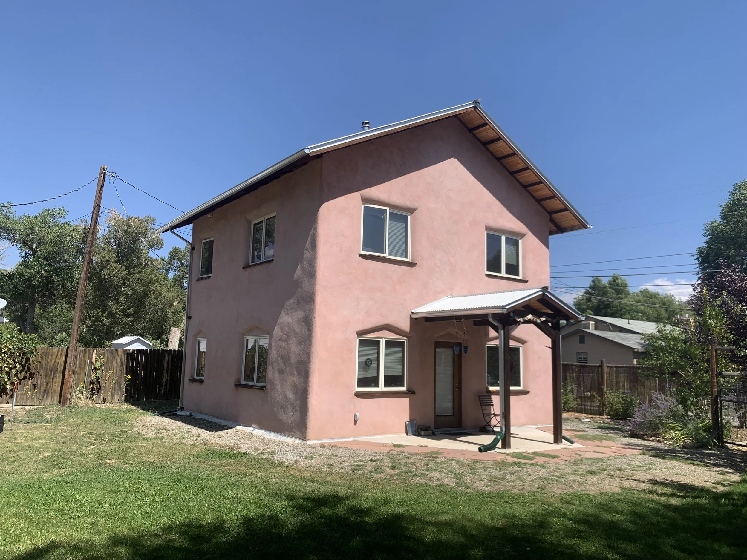 A pink, two-story house with white-framed windows and a small covered patio area in the backyard. There is a grassy lawn, a wooden fence, and trees surrounding the house, with a clear blue sky above.