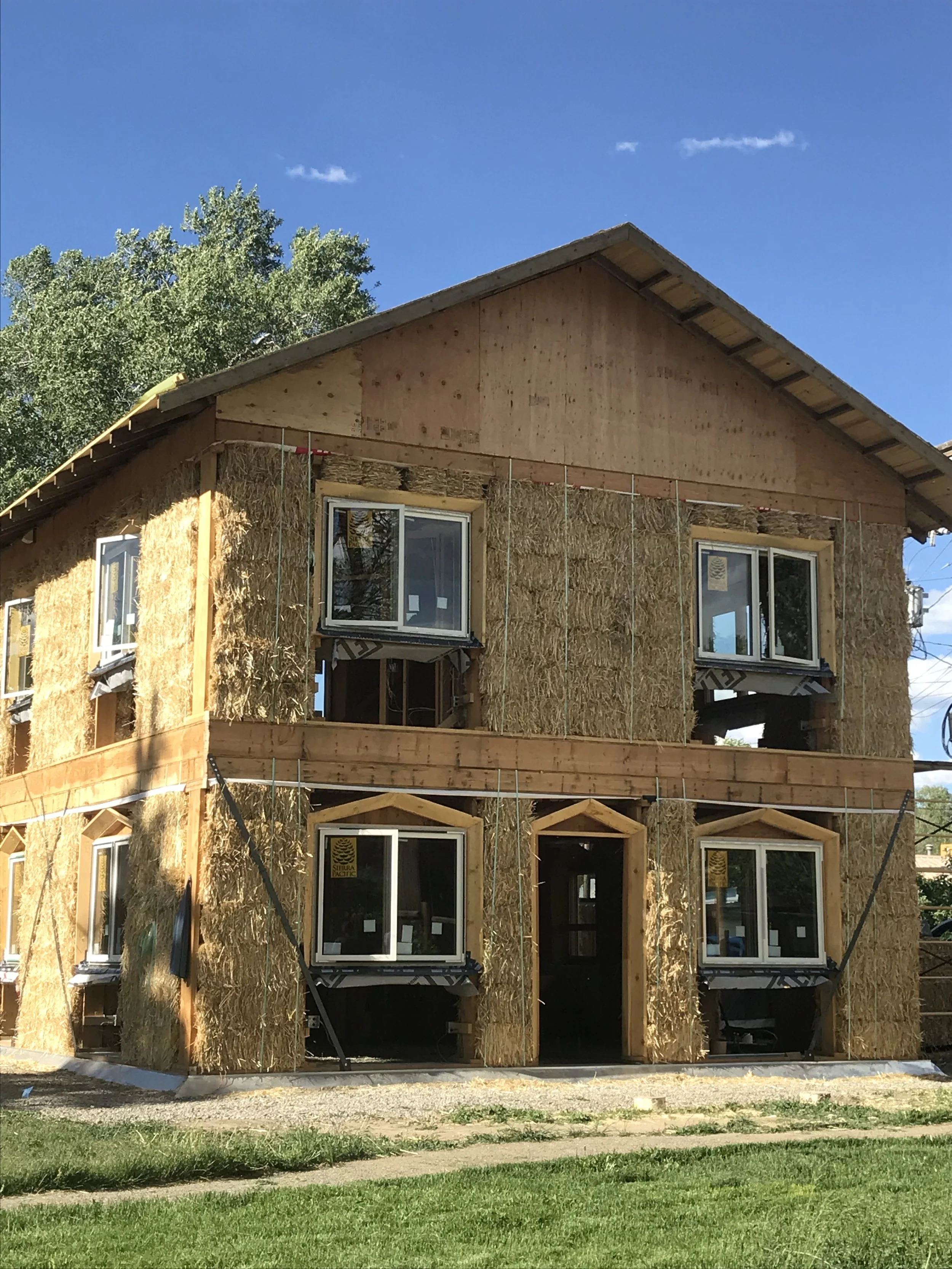 A house under construction with wooden framing and insulation, with a clear blue sky and trees in the background.