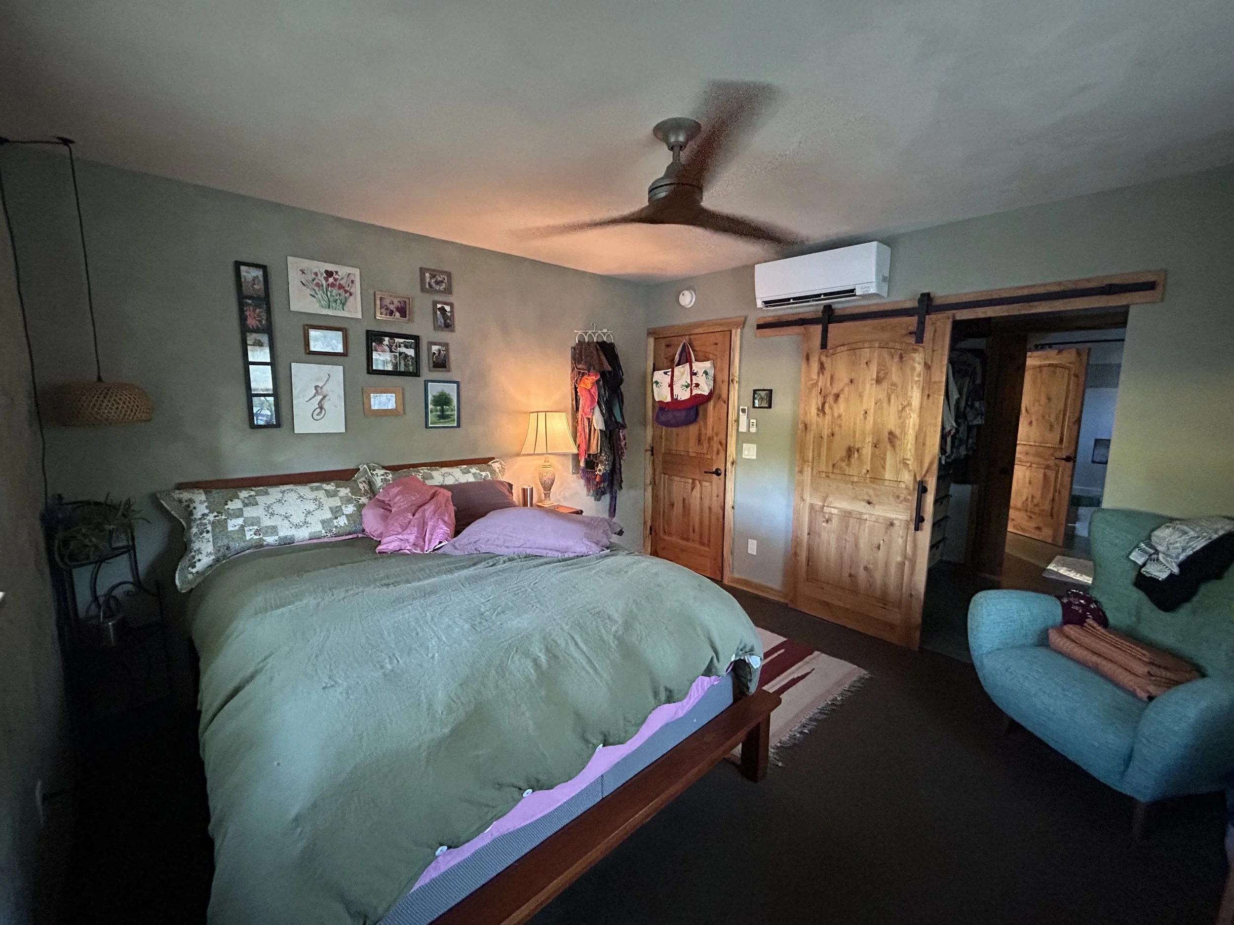 A bedroom with a large bed covered in a green quilt, pink pillows, and a quilted bedspread. There's a collage of framed photos and artwork on the wall above the bed, a wooden nightstand with a lamp, and a sliding barn door leading to an adjoining roo