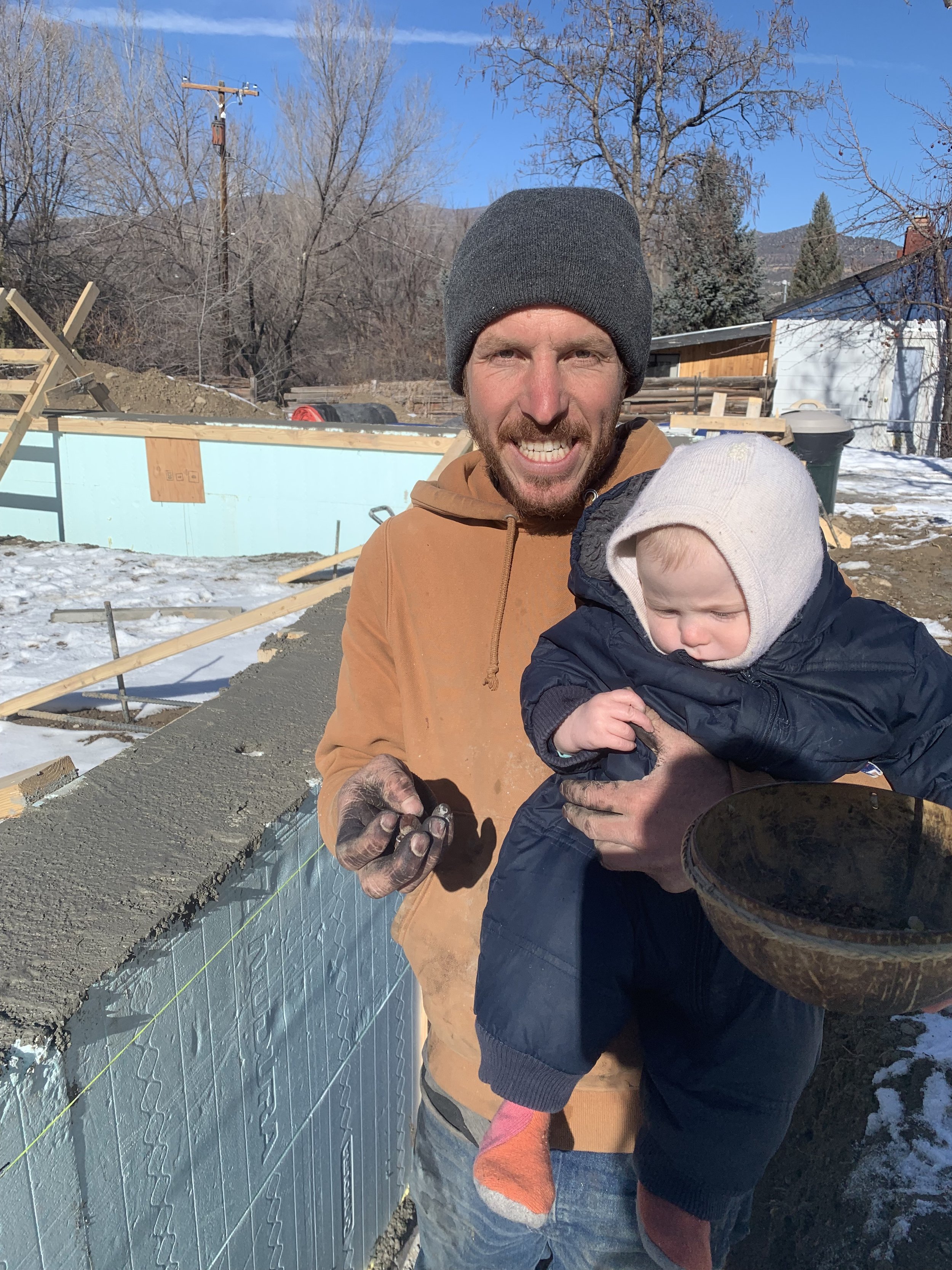 Two people, a man and a child, are smiling and working together on a construction site outdoors in winter. The man is wearing a gray beanie and a tan hoodie, and the child is wearing a white hat and a dark jacket, with snow on the ground and a partly cloudy blue sky in the background.