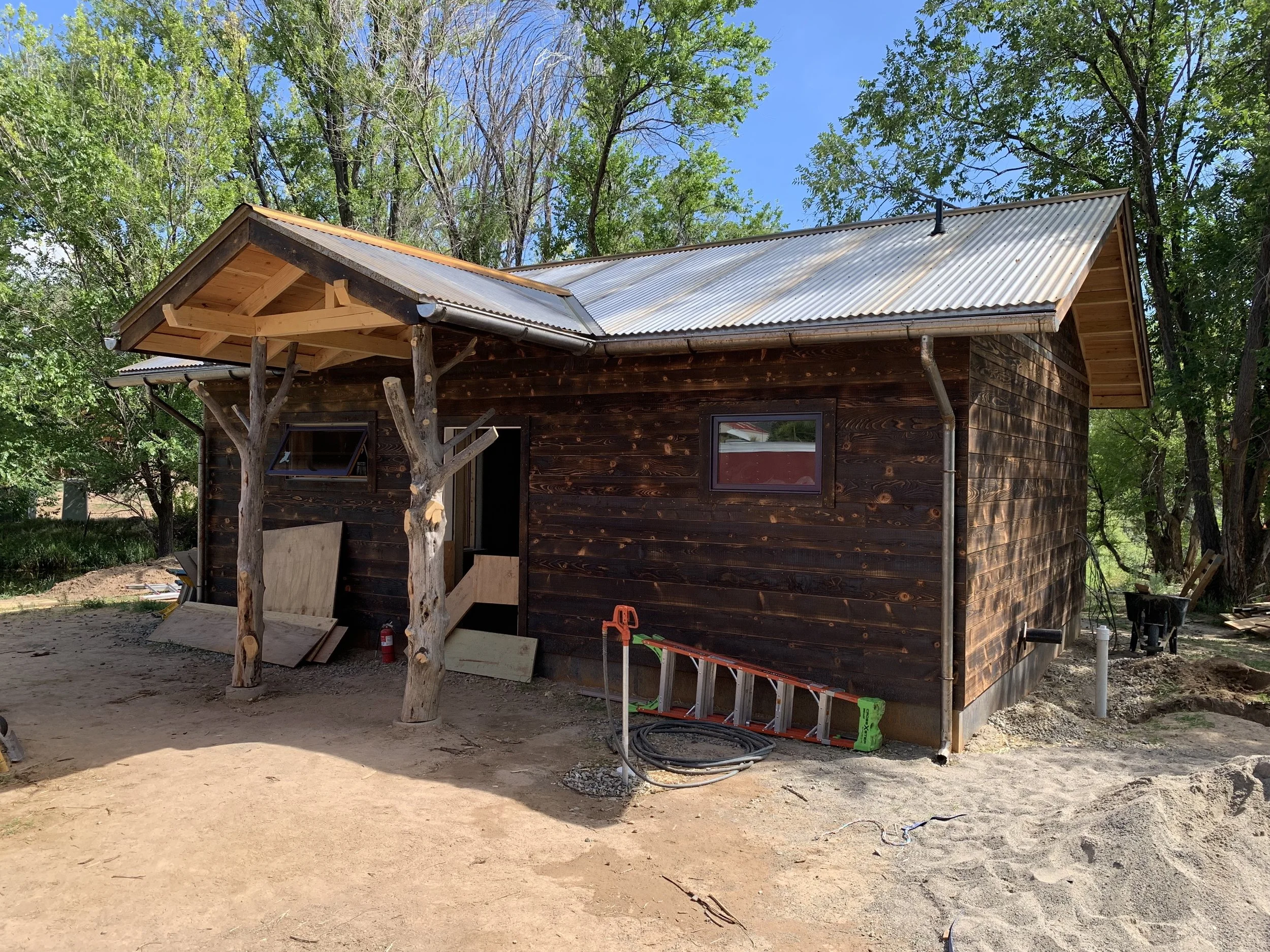 A small wooden house under construction with a metal roof, surrounded by green trees and a dirt ground, with construction tools and materials nearby.