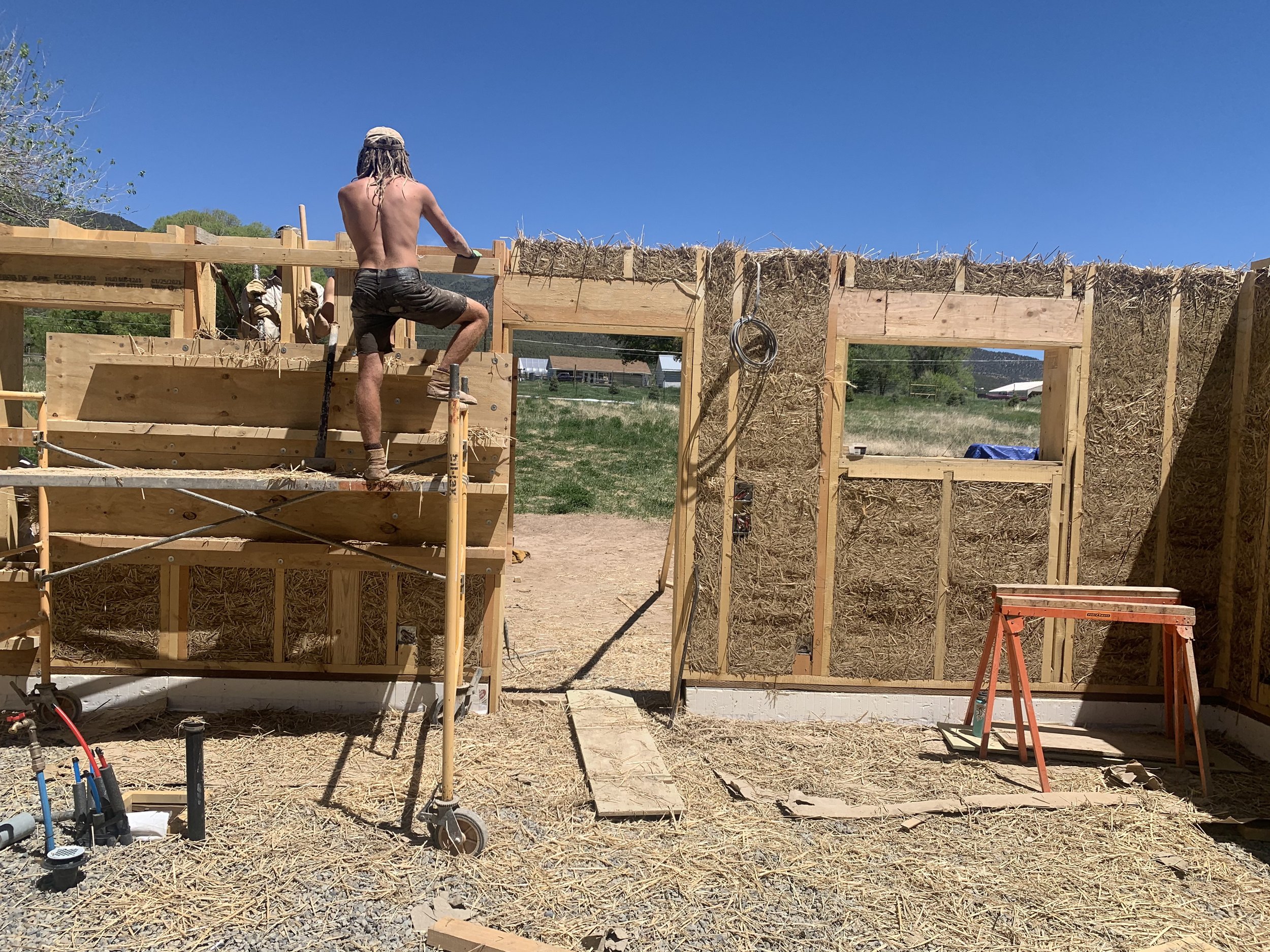 A construction site with wooden framing and straw insulation being installed, a shirtless man with dreadlocks working, and a blue sky with some trees and buildings in the background.