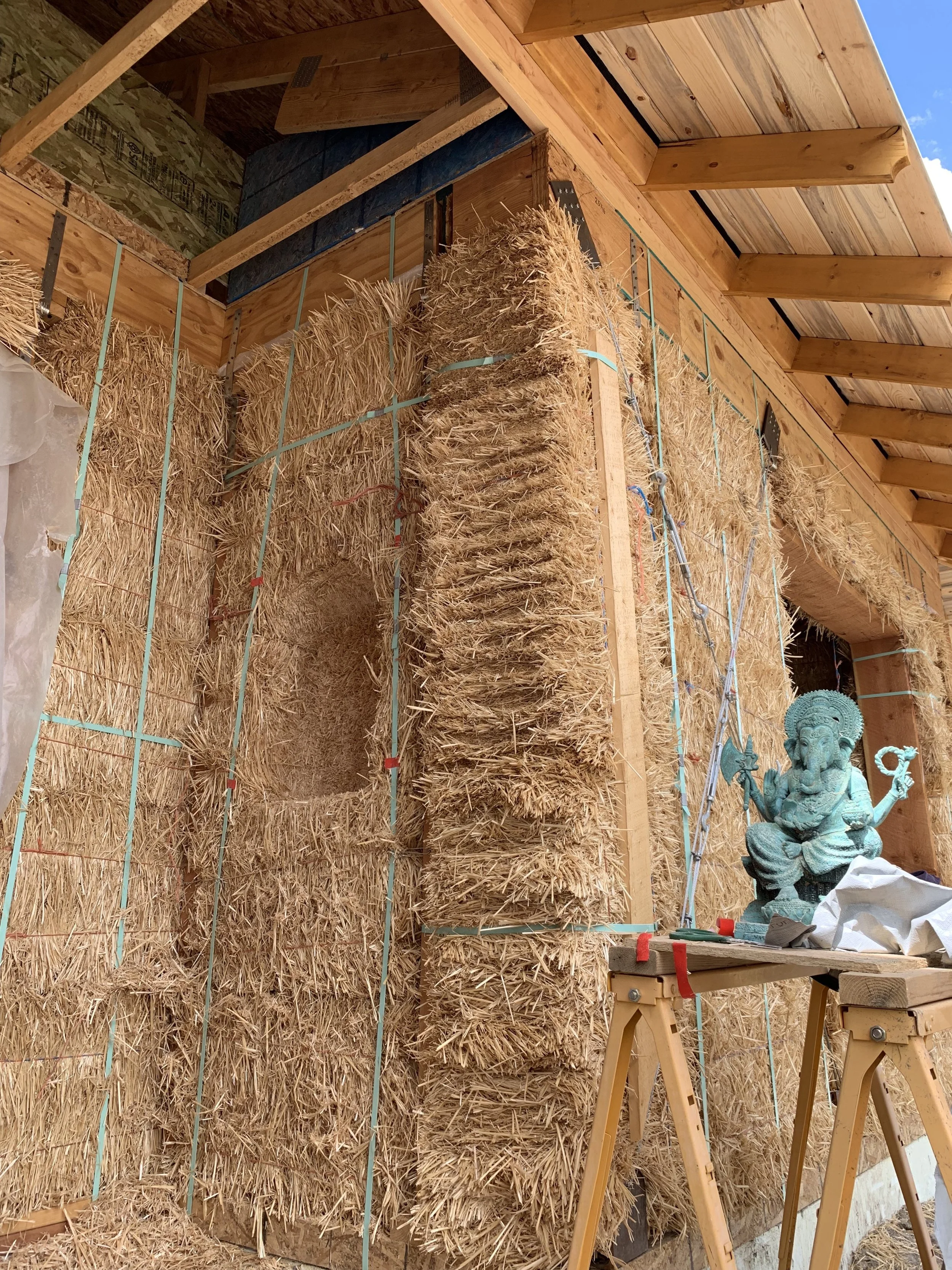 Construction site with straw bales stacked inside a wooden frame, a statue of Ganesha on a work table, and construction tools nearby, under a wooden roof with a view of blue sky.