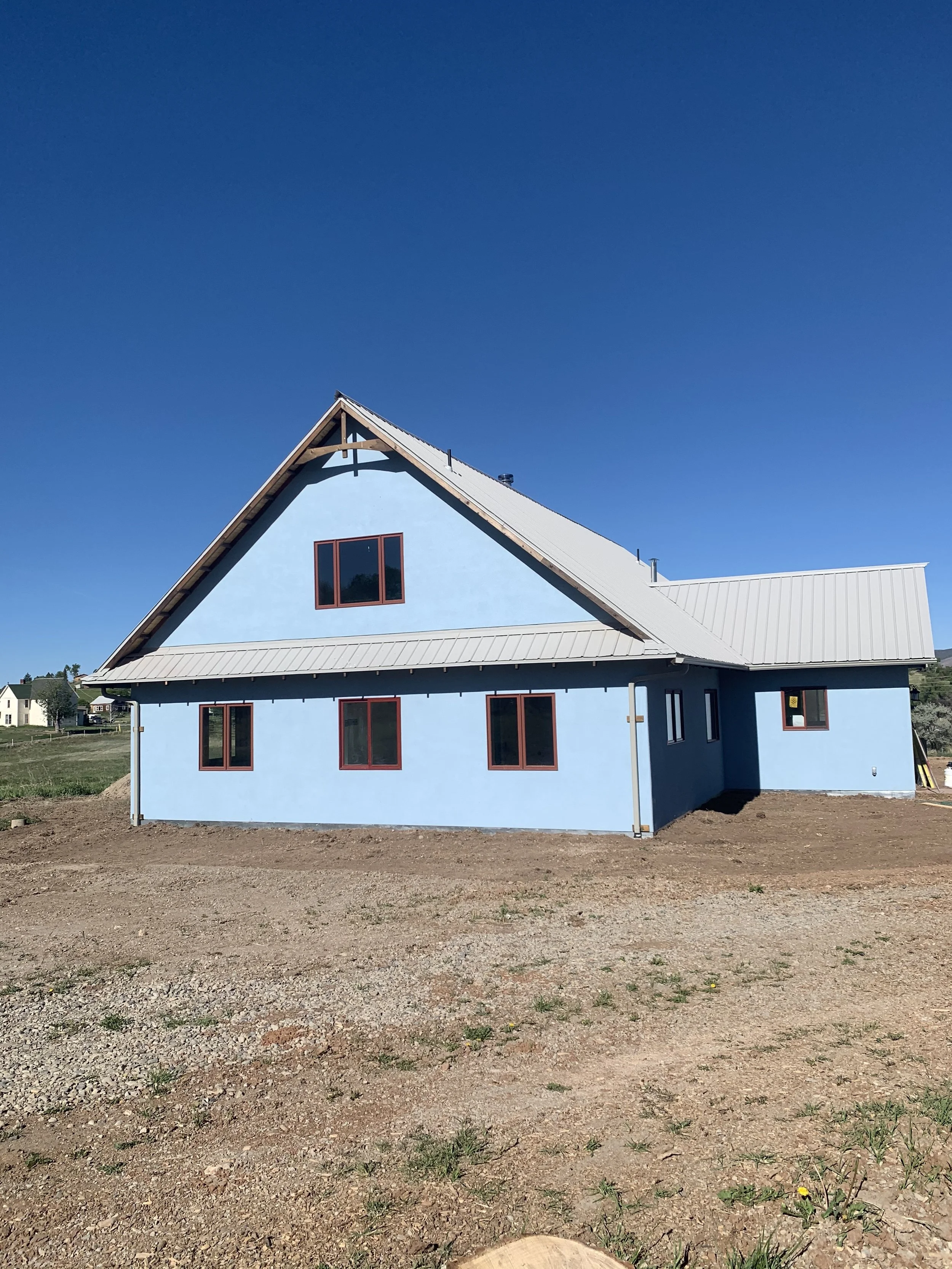 A blue house under construction with a steep roof, multiple windows, and a large open area around it on a clear day.