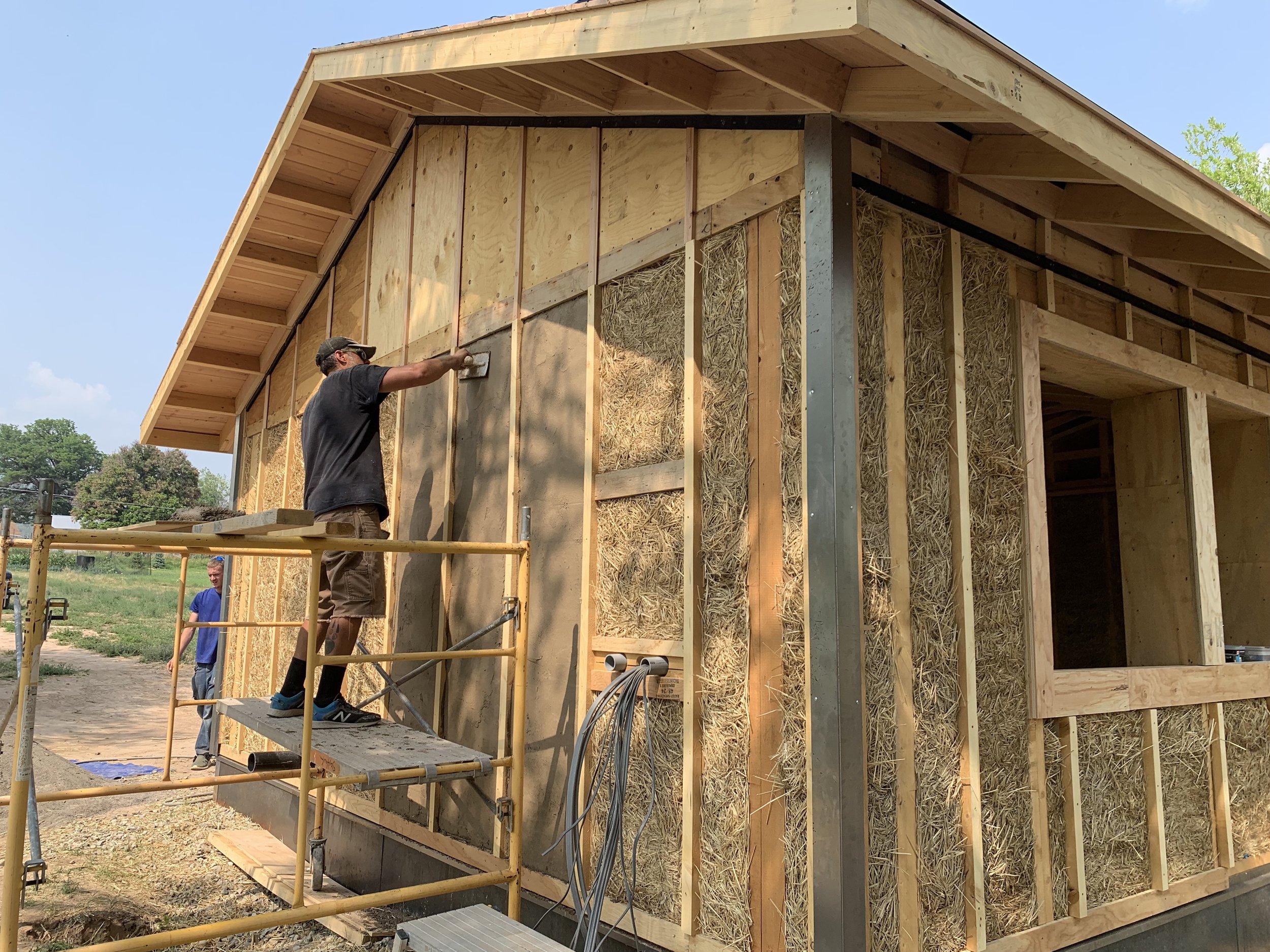 A construction worker is applying stucco to the exterior of a framed building under construction. The building has foam board insulation and straw-filled wall sections, with a roof framework above.