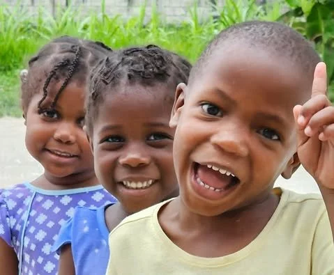 Three smiling children outdoors, with a green background, one of them raising a finger.
