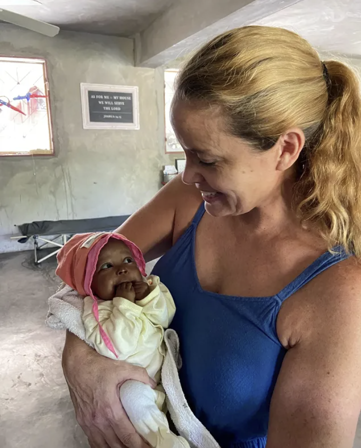 A woman with blonde hair in a ponytail holding a baby girl with a pink hat and yellow outfit, smiling at each other inside a simple room with a gray wall and windows.