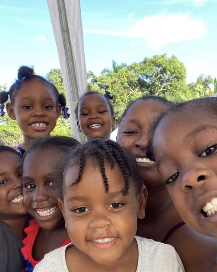 Group of smiling children taking a selfie outside under a canopy with trees in the background.