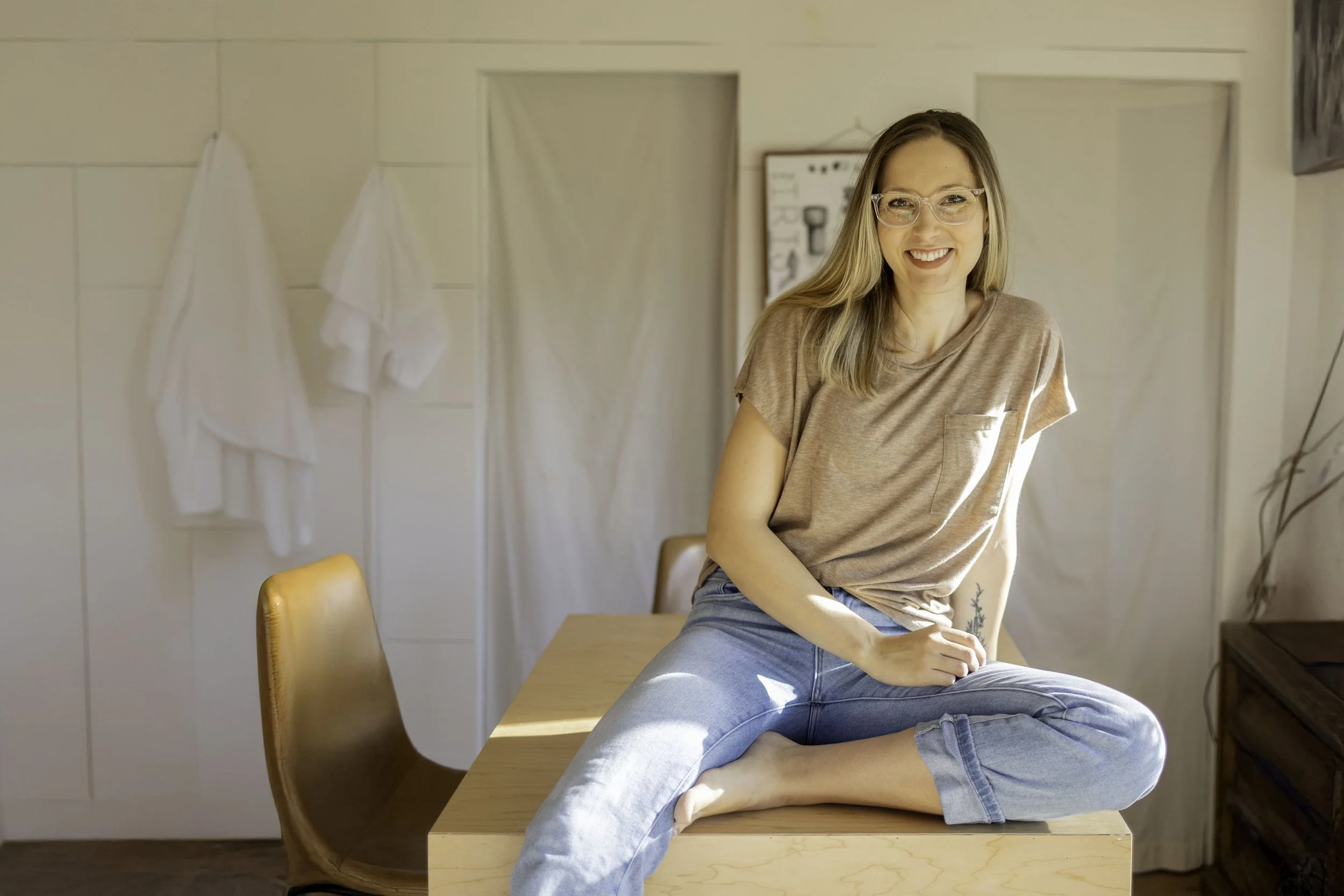 A woman with long blonde hair and glasses, sitting on a kitchen counter with her legs crossed, smiling at the camera.