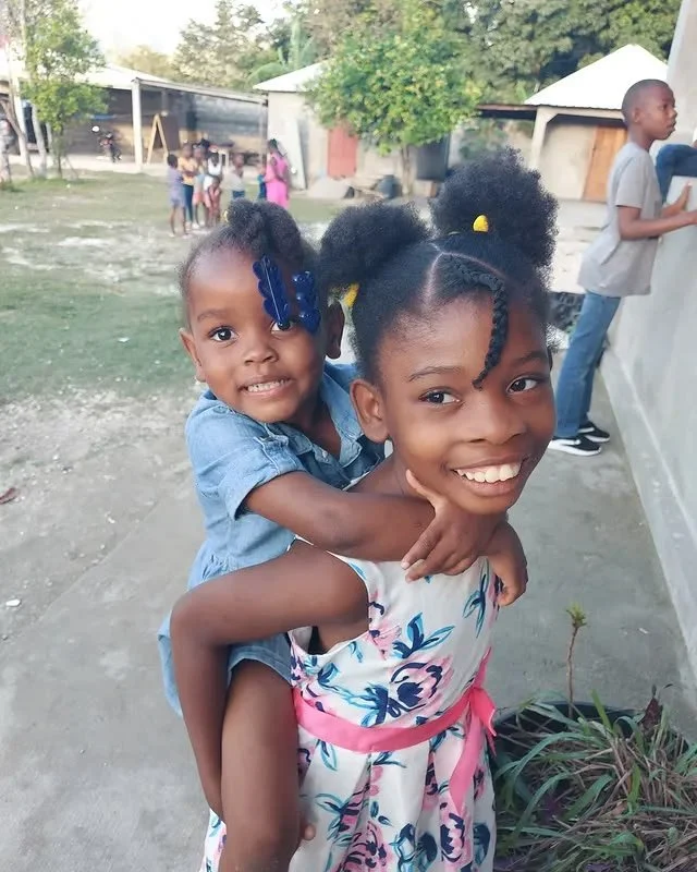 Smiling young woman with braided hair and a pink and white floral dress giving a piggyback ride to a young girl with puffy hair and a denim dress, in an outdoor setting with several other children in the background.