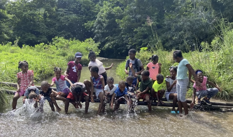 Children playing and splashing in a river with lush green trees in the background.