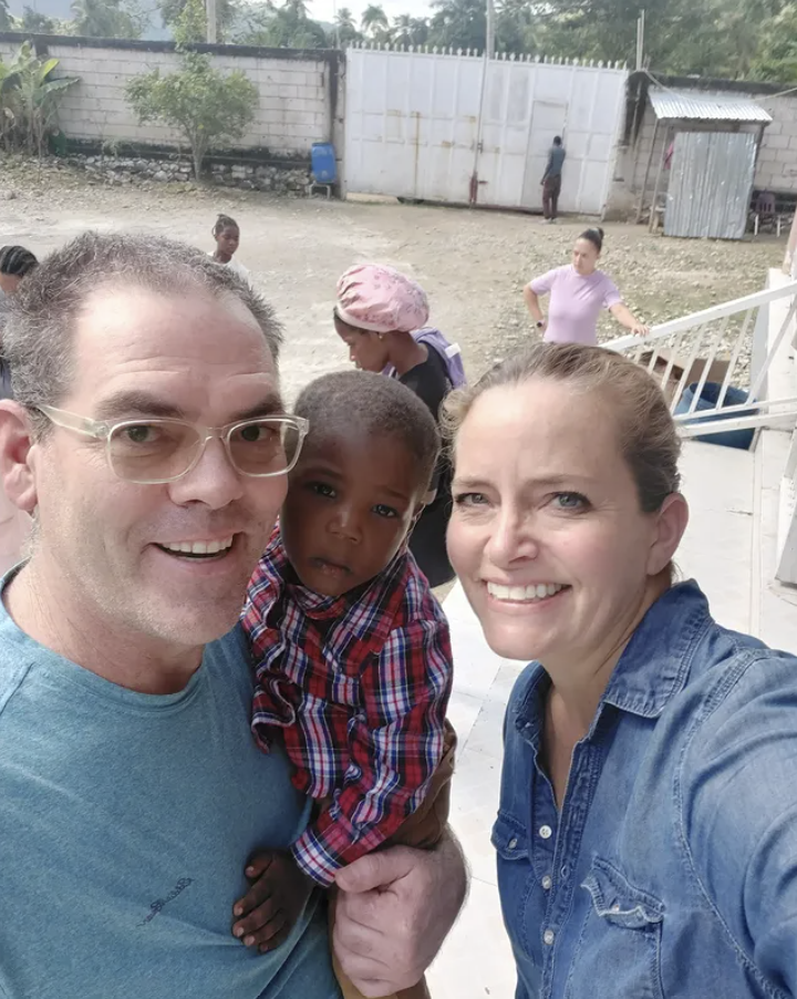 A group of four people, including two adults and a young boy, taking a selfie outdoors. The man is wearing glasses and a blue shirt, the woman has brown hair tied back and is wearing a denim shirt, and the boy is in a red and blue plaid shirt. In the background, there are two women, one wearing a pink hat and another in a pink shirt, along with a few other people and a white fence.