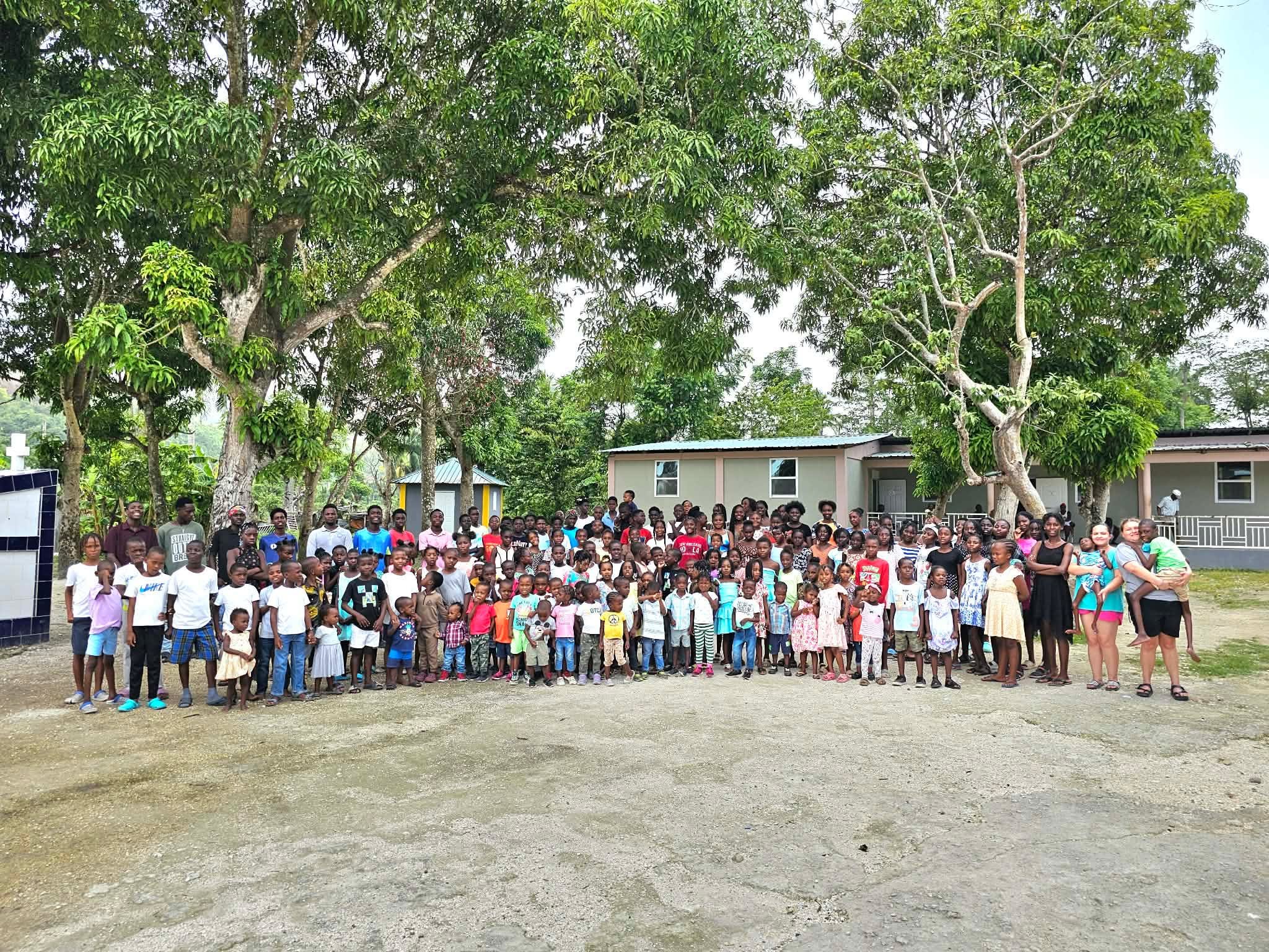 A large group of children and adults gathered outdoors in front of trees and a building, posing for a group photo.