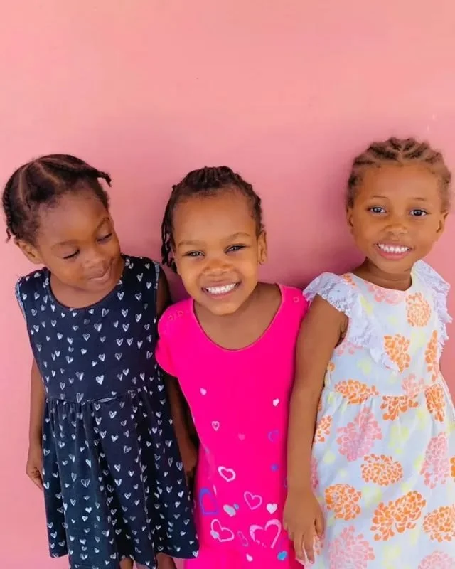 Three young girls standing against a pink wall, smiling and wearing colorful dresses.