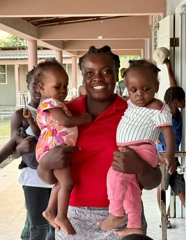 A smiling woman in a red shirt holding two young girls, one in each arm, with children and people in the background in an outdoor setting.
