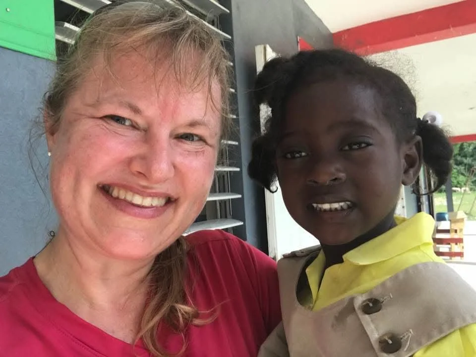 A smiling woman with light skin and a young girl with dark skin standing together in an outdoor setting, possibly at a school or community area. The woman is wearing a red shirt and the girl is in a yellow and beige school uniform.