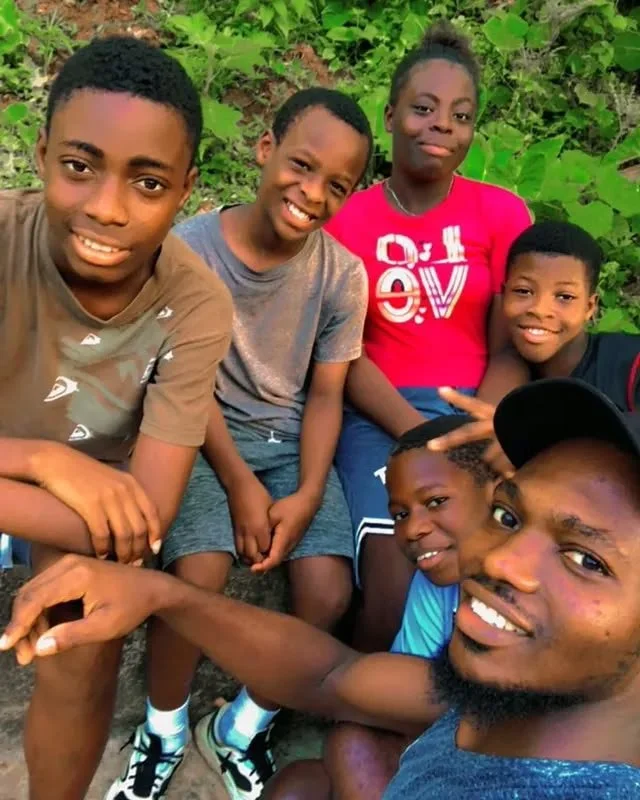 A group of seven children smiling outdoors among green foliage.