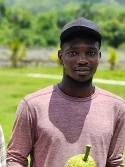 Young man with dark skin, wearing a black cap and a light purple long-sleeved shirt, standing outdoors with green trees and grass in the background.