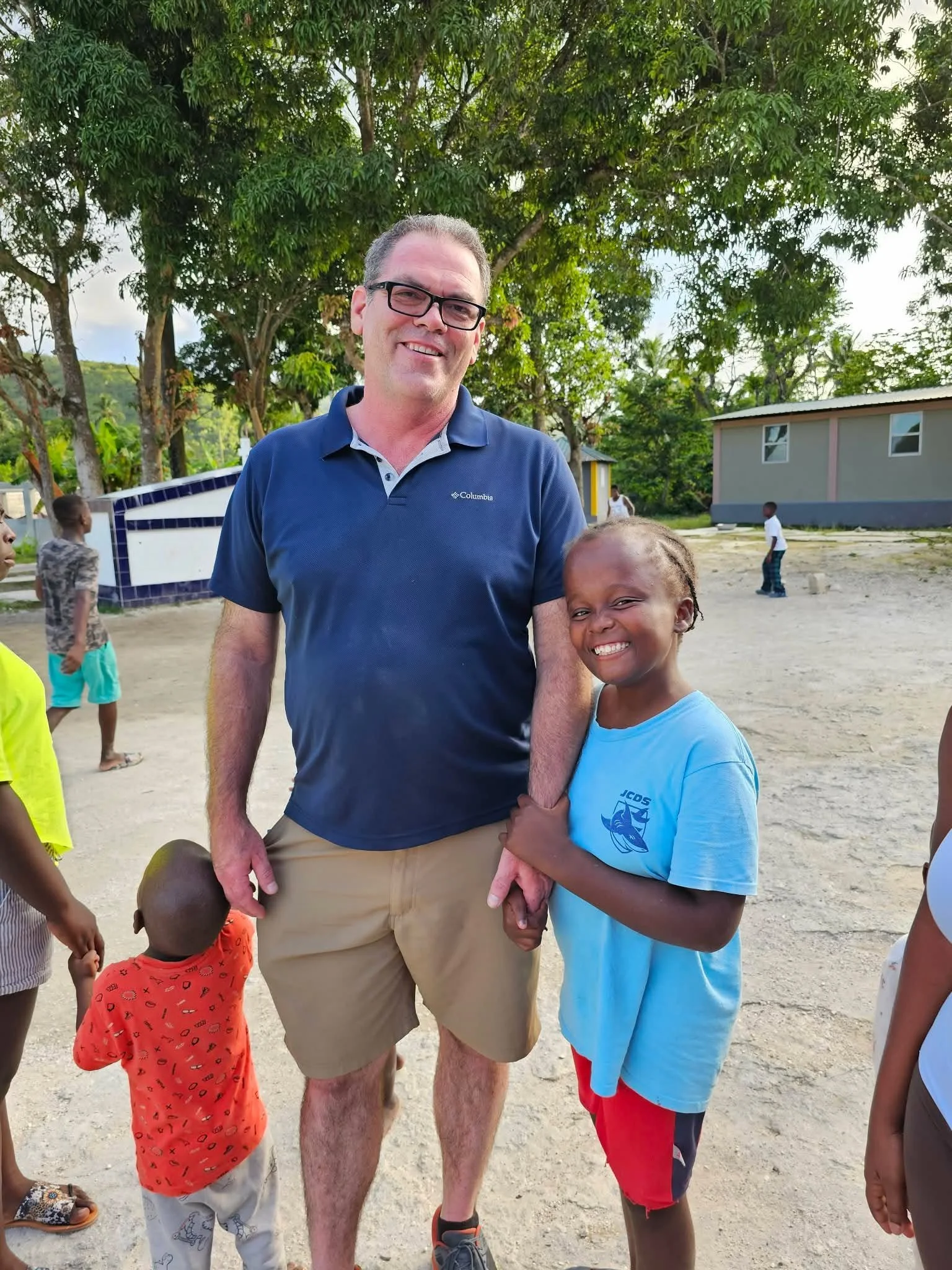 Man and children standing outside in a sunny area with trees and a small building in the background. The man is wearing glasses and a blue polo shirt, smiling, holding a basketball. The children are smiling, one wearing a light blue shirt and red shorts, others in colorful clothing.