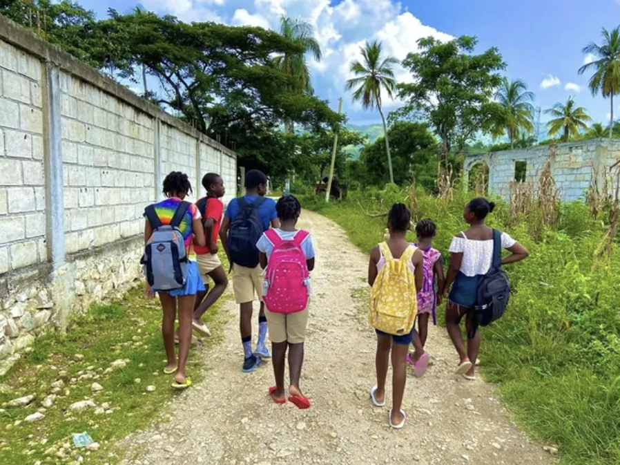 Group of children walking down a dirt road in a rural area with palm trees, green vegetation, and partially built houses.