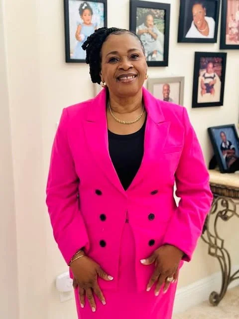 A smiling woman in a pink blazer and black top standing indoors in front of framed photos on a wall.