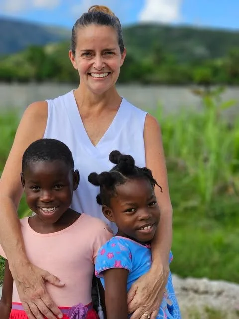 A smiling woman standing outdoors with two young girls, all three are close together and smiling, with green trees and a lake in the background.