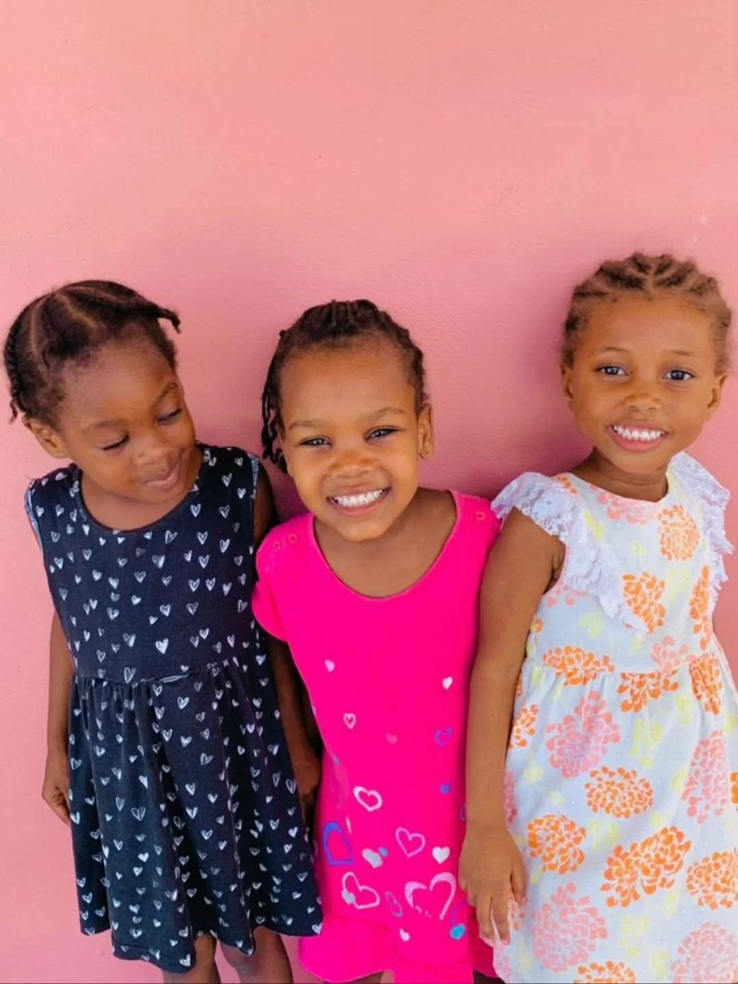 Three young girls standing against a pink wall, smiling and wearing colorful dresses.