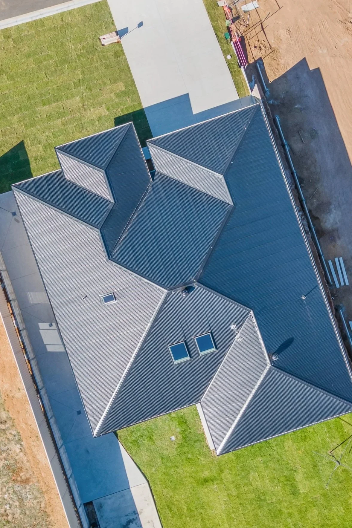 Aerial view of a modern house with a large gray metal roof, small skylights, and a surrounding lawn with paved pathways.
