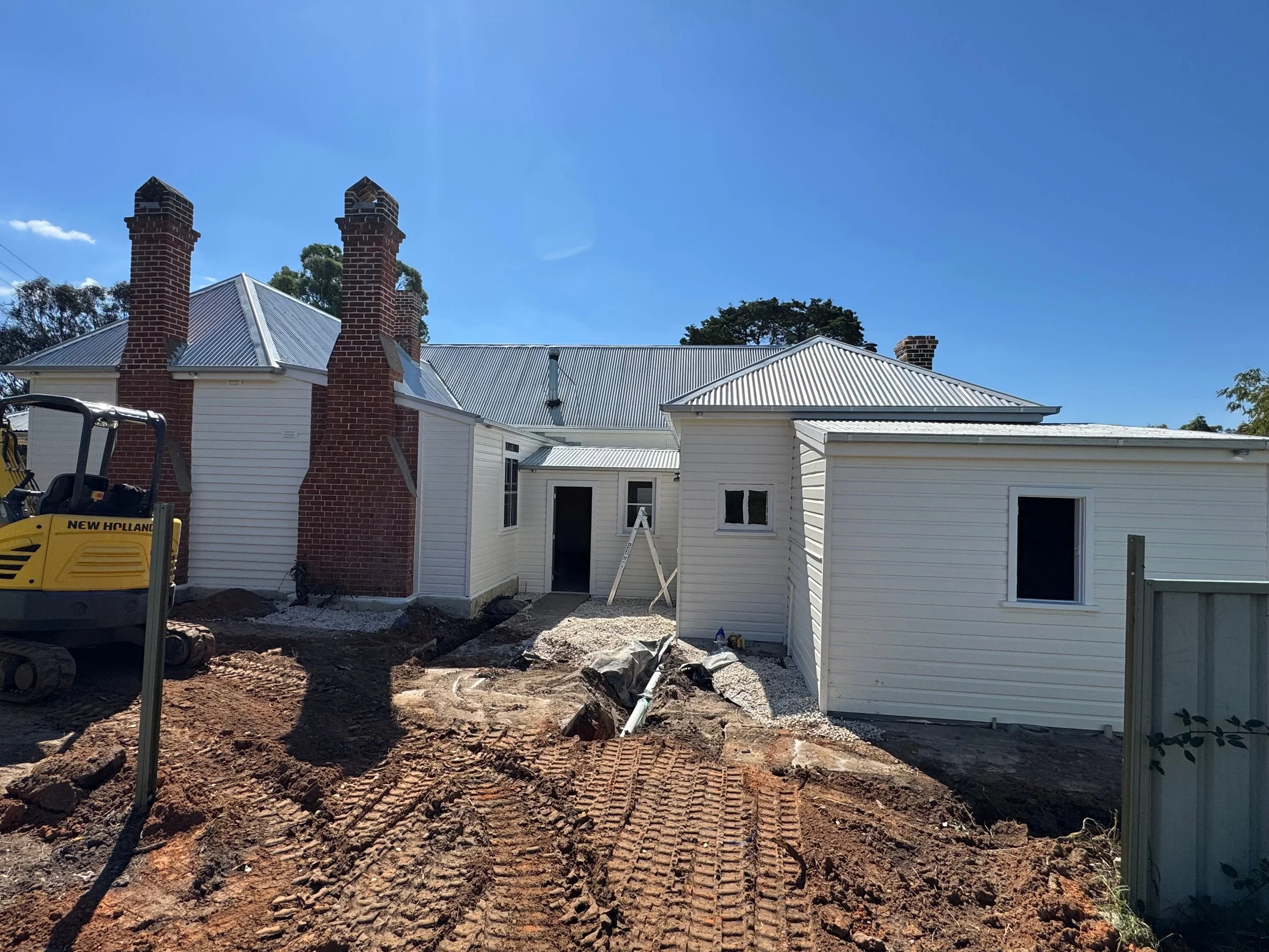 Construction site with a white house, brick chimneys, and a partially excavated yard with construction equipment and dirt.