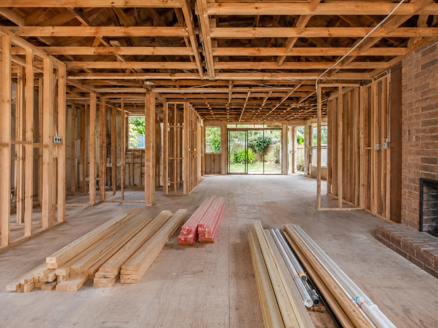 Interior of a house under construction, showing exposed wooden framing, construction materials, and a view of the backyard through large glass doors.