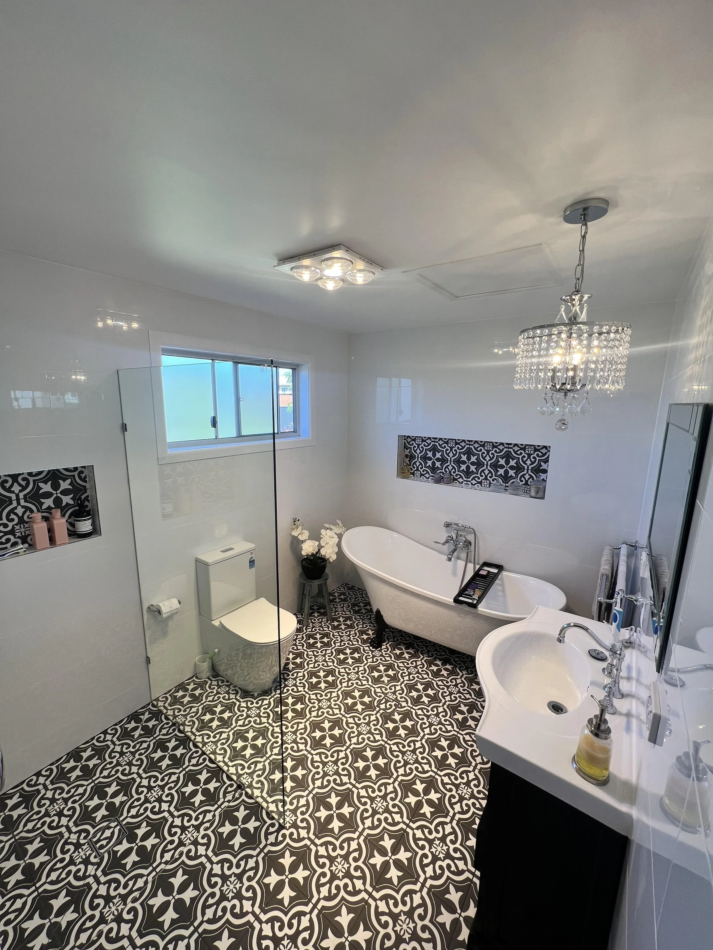 A modern bathroom featuring patterned black and white floor tiles, a white clawfoot bathtub, a white sink with a black vanity, a crystal chandelier, a window, and a toilet. Decorations include a potted white flower and various toiletries.