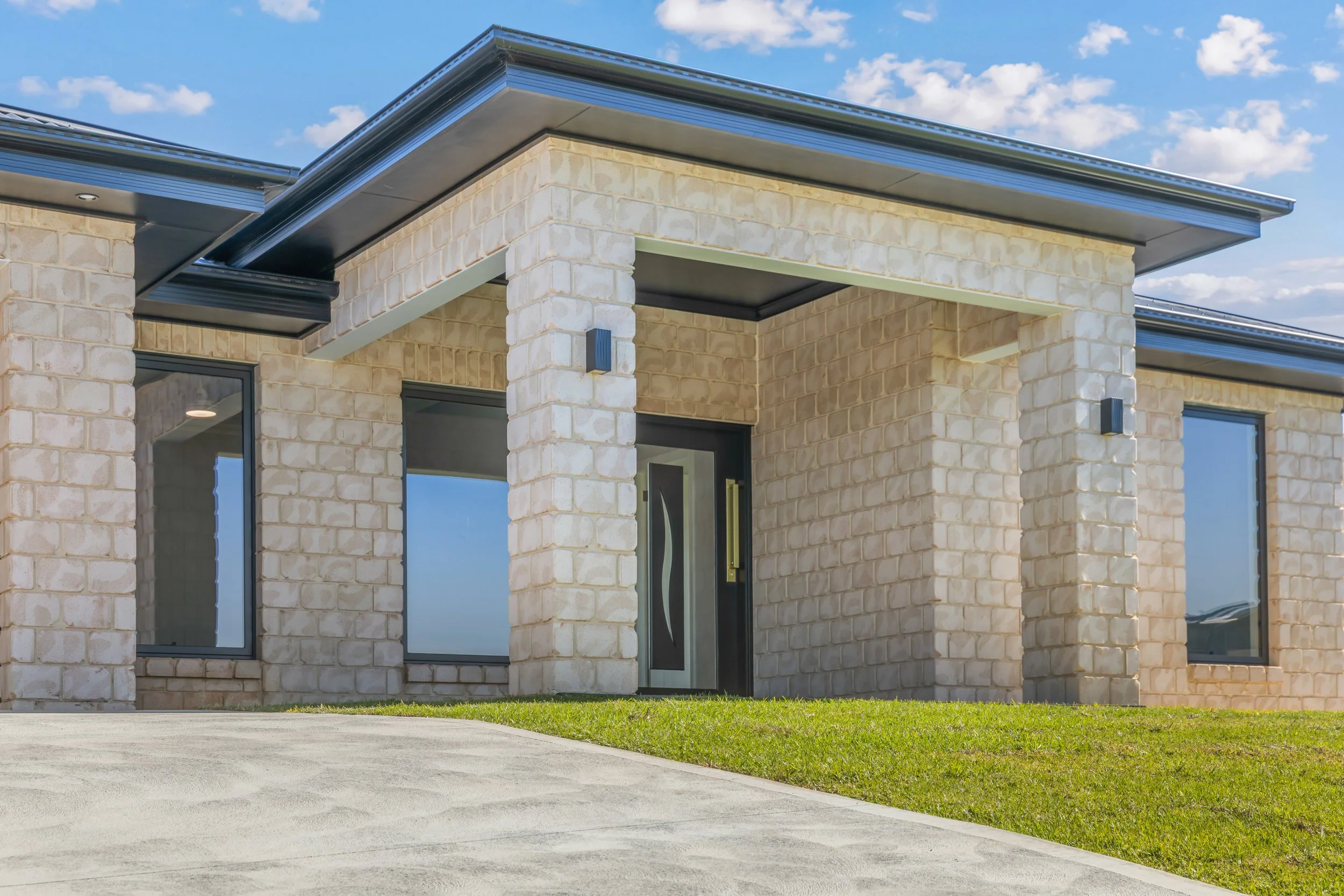 Front view of a modern house with brick walls, large glass windows, and a black metal roof under a partly cloudy sky.