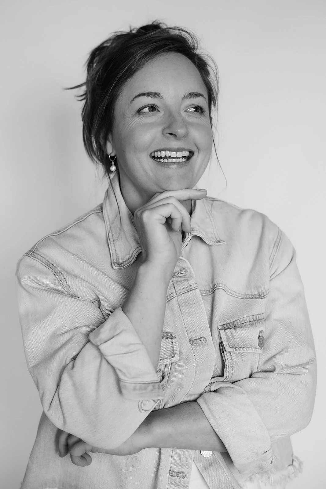 Black and white portrait of a woman smiling with hand on chin, wearing a denim jacket, earrings, and short hair.