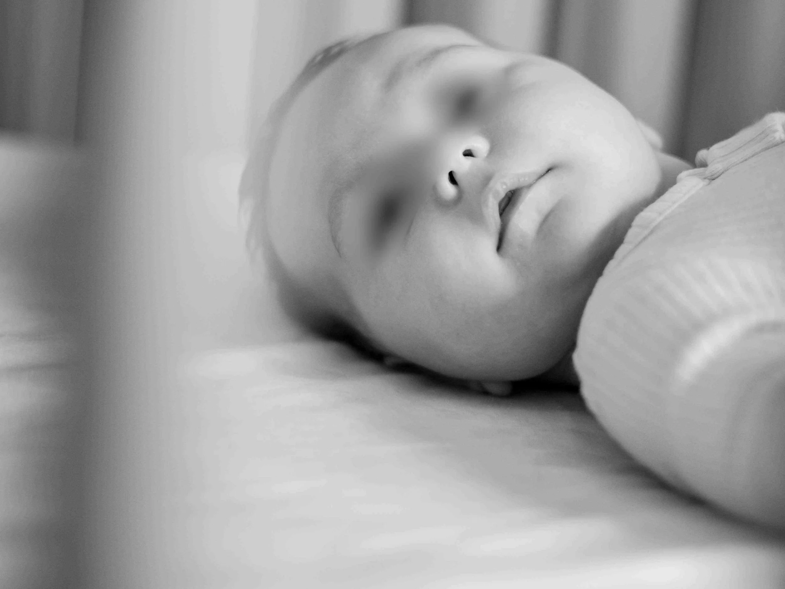 A close-up black and white photograph of a baby with a hand touching their lips, lying down on a soft surface.