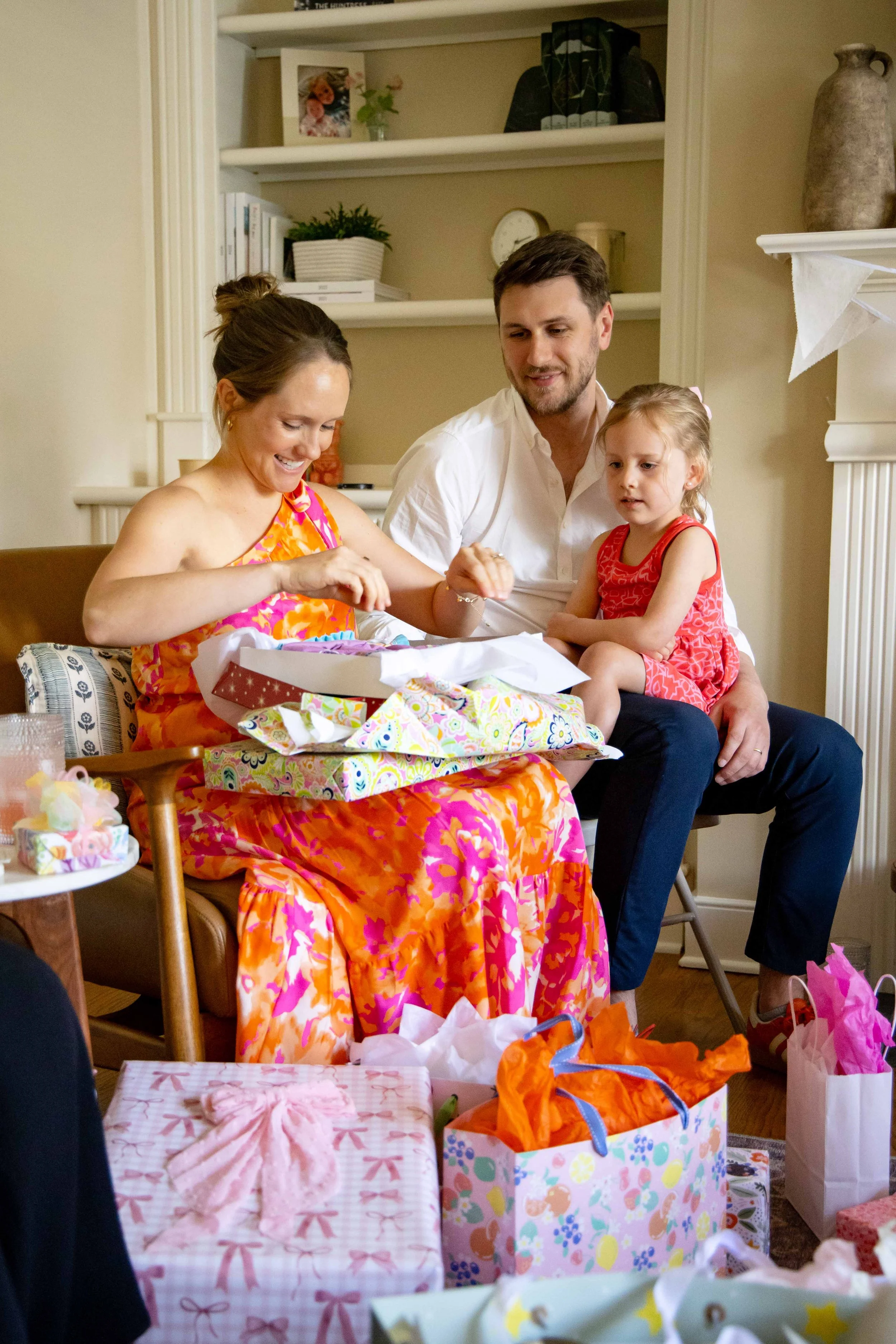 A woman in a colorful dress and a man with a girl sit together during a gift opening event with presents and wrapping paper around them.